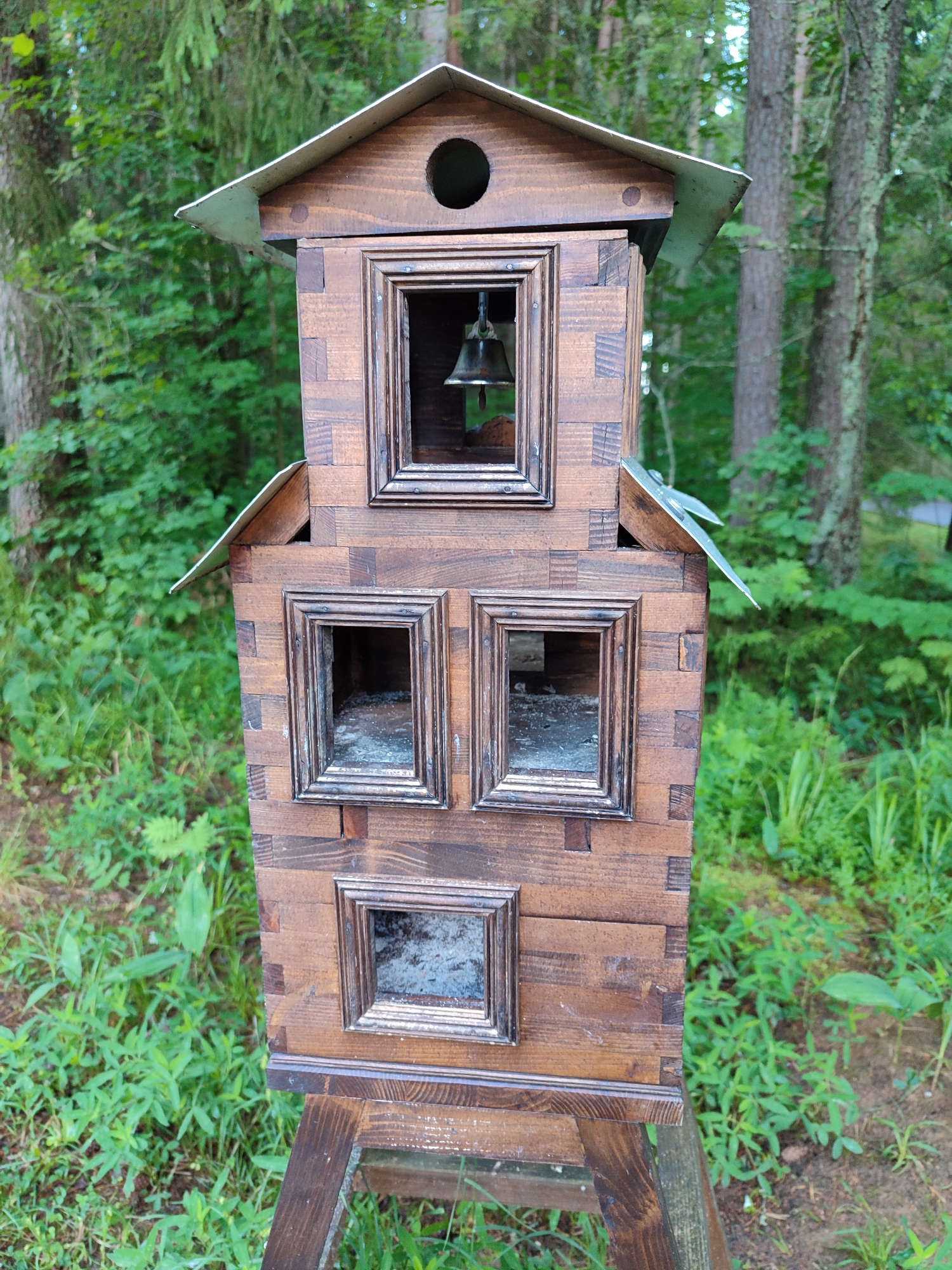 A tall, wooden, multi-tiered structure with a metal roof stands outdoors in a natural setting. The structure has several square openings framed with dark wood, some of which appear to be empty, while one at the top contains a small bell. The wood of the structure is comprised of many small, interlocking rectangular pieces, giving it a textured appearance. It is supported by a simple wooden base. The background consists of green foliage and trees, suggesting a park or wooded area. The lighting is natural, indicating daylight. There are no people or animals visible in the image.