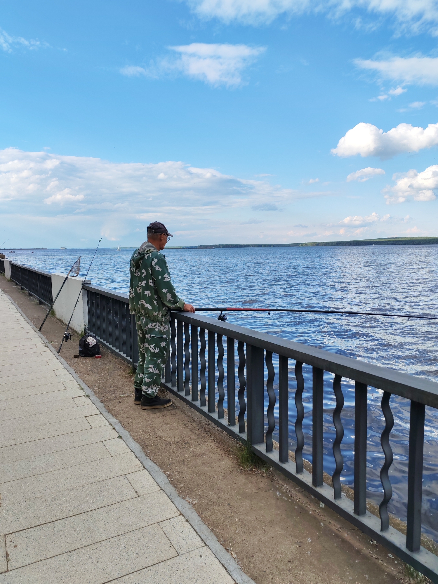 A man in camouflage clothing is fishing on a paved embankment beside a wide, blue river under a partly cloudy sky. He is wearing a cap and glasses and is holding a fishing rod. Two other fishing rods are propped up on the embankment behind him, along with a black bag and a fishing net. The river is calm with gentle ripples, and distant sailboats and a large vessel are visible on the water. A treeline borders the far bank of the river. The scene appears to be a pleasant, casual afternoon for fishing.