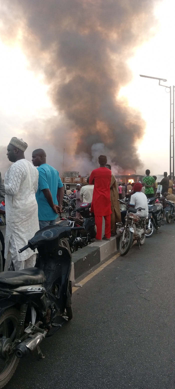 A group of people stand near a road, observing a fire and plumes of smoke. Parked motorcycles line the road, positioned on the curb. Some individuals are dressed in traditional attire, while others wear more casual clothing. The smoke rises high, obscuring the details of the building where the fire appears to be located. A streetlight pole is visible on the right edge. The overall scene indicates a serious event drawing the attention of onlookers.