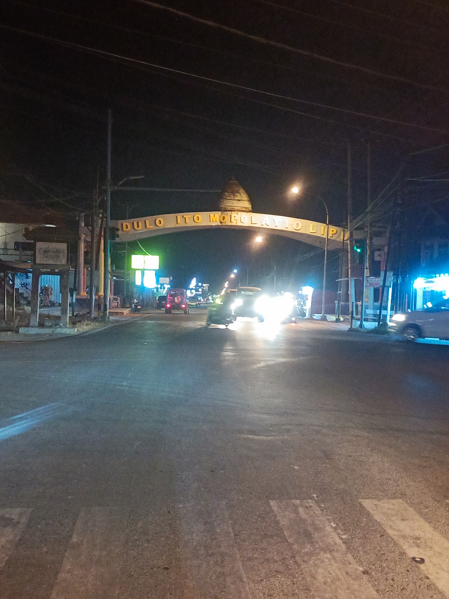 An archway with illuminated text spans across a road at night. The text on the archway reads "DULO ITO MOHOLAYO LIP". A dome-like structure sits atop the archway. The scene depicts a street in Pentadio Barat, Indonesia, with traffic lights, streetlights, and various vehicles on the road. Some buildings and structures line the sides of the road. The overall impression is that of a town entrance or landmark at night.