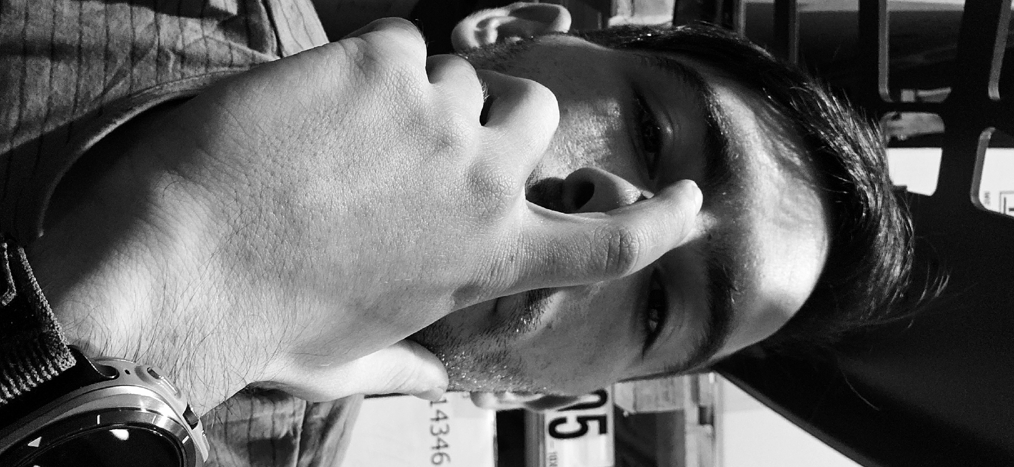 The image is a close-up, black-and-white photograph of a man, potentially in Plovdiv, Bulgaria. He is shown from the chest up, turned sideways. His left hand is raised to his face, with his fingers near his nose and mouth. He is wearing a striped shirt and a watch on his wrist. Behind him, partially obscured, is a sign with numbers "14346" and "75" visible. A metal structure is also visible in the background.