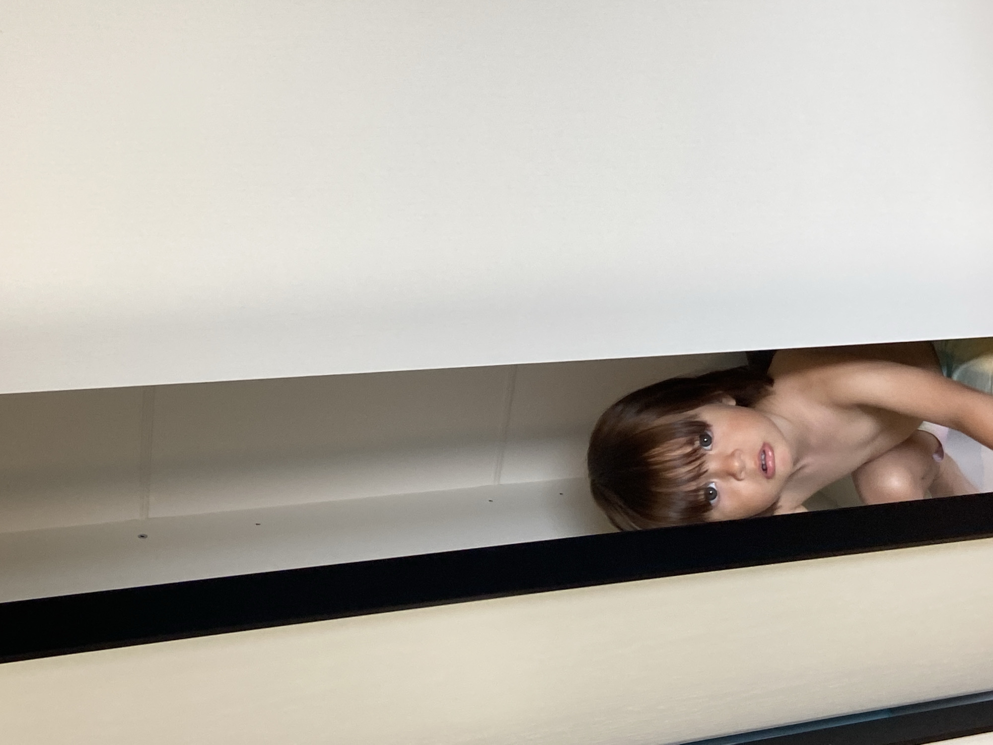 A low-angle, eye-level shot captures a young child's face peering out from what appears to be a partially open drawer or cabinet in Tyumen, Russia. The child, with shoulder-length brown hair and bangs, is looking directly at the camera with wide eyes and a slightly open mouth, giving an impression of curiosity or playful mischief. Their bare shoulders and part of their torso are visible, suggesting they are without a shirt. The upper half of the image is dominated by the light-colored underside of what seems to be another drawer or shelf, creating a narrow gap through which the child is looking. Below the child's face, a thick black horizontal bar separates the immediate foreground from the background, which is the interior of the drawer/cabinet where the child is situated. The lighting is soft and even, without harsh shadows, suggesting an indoor setting. The scene evokes a playful "hide-and-seek" moment. No specific time of day or weather can be discerned, and there is no visible text.