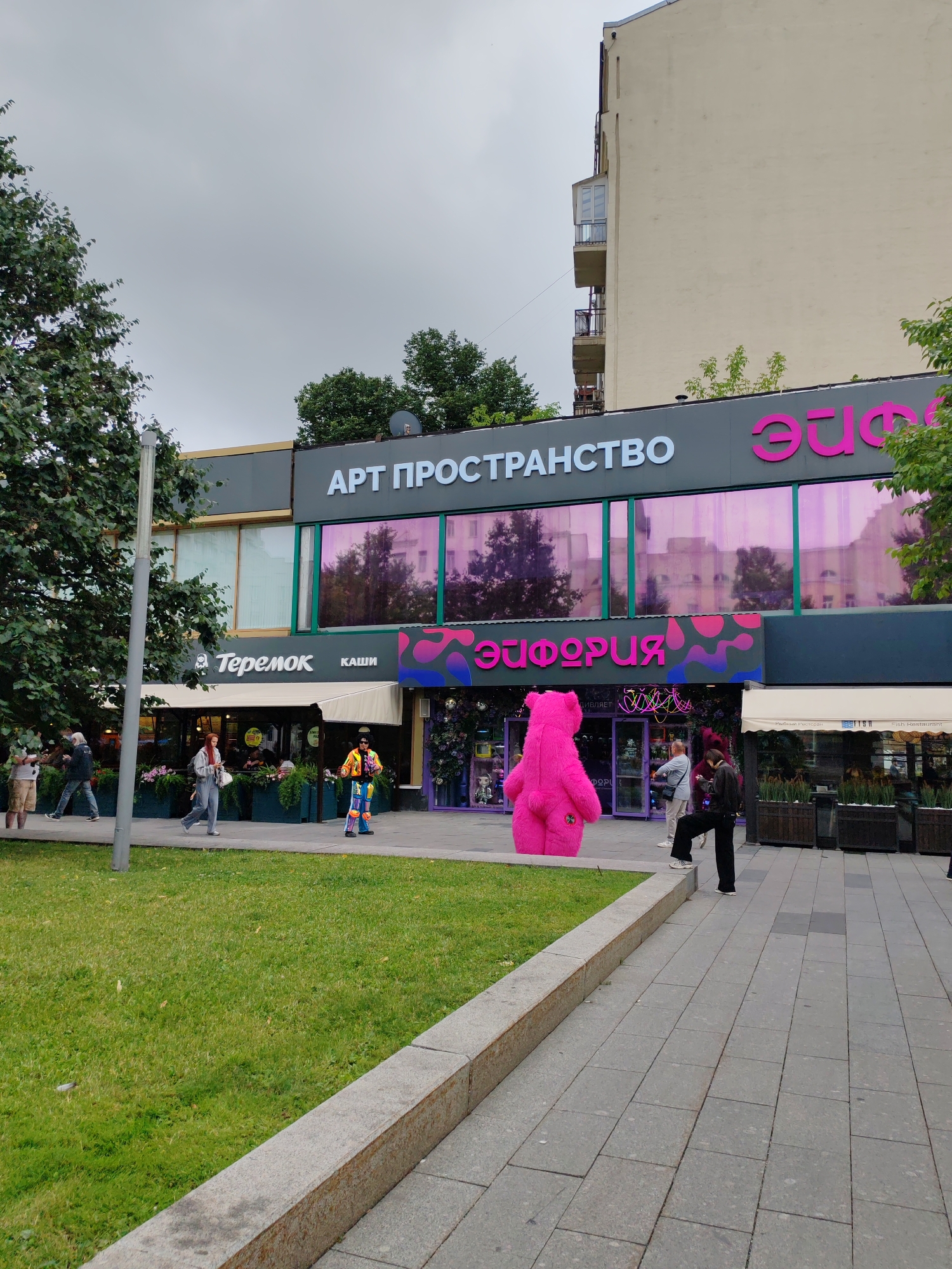 The image depicts a street scene in Moscow, Russia, in front of a commercial establishment. The building features large windows with a pink, reflective tint and prominent signage that reads "АРТ ПРОСТРАНСТВО" (Art Space) and "ЭЙФОРИЯ" (Euphoria). Below the main signage, there is a sign for "Теремок КАШИ" (Teremok Kashi), a fast-food chain specializing in kasha (porridge).
In the foreground, a large, bright pink inflatable bear stands near the entrance of the "ЭЙФОРИЯ" establishment, serving as a visually striking attraction. Several people are present in the scene: a person in a colorful, patterned outfit stands near the pink bear, appearing to be an entertainer or mascot; others walk along the sidewalk in various directions. One individual is seen walking away from the camera, wearing a white hooded jacket and blue jeans. Another person, a woman with red hair, walks towards the camera, carrying a white bag. Two individuals stand near the entrance of "ЭЙФОРИЯ," one looking at their phone.
The environment includes a well-maintained lawn with trees on the left side of the image and a paved sidewalk. The sky is overcast, suggesting cloudy weather. The architectural style of the adjacent building in the background is typical of urban Russian apartment blocks, with balconies and multiple floors. The overall atmosphere appears to be a bustling urban setting with a focus on commercial activity and public spaces.