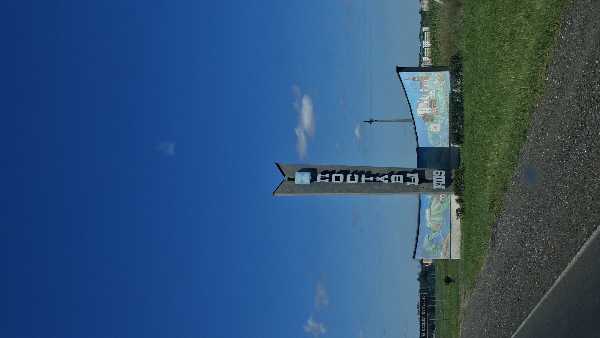 The image shows a monument at the entrance to the city of Postavy, Belarus. The monument is a tall, grey, concrete structure with the city name "ПОСТАВЫ" written vertically in blue letters. Below the name, there is a blue shield with a white symbol. To the right of the monument, there is a curved mural depicting scenes of the city and its surroundings, including buildings, a river, and fields. In the foreground, there is a paved road and green grass. In the background, there are trees and buildings, suggesting the city is nearby. The sky is blue with a few scattered clouds. The monument also displays the year "1409" in white numbers. On the left side of the image, a road sign is visible that reads "Касце́л XVII ст. 20", indicating a church from the 17th century located 20 kilometers away. The overall scene is an entrance marker to a Belarusian city, likely taken from a vehicle driving past.