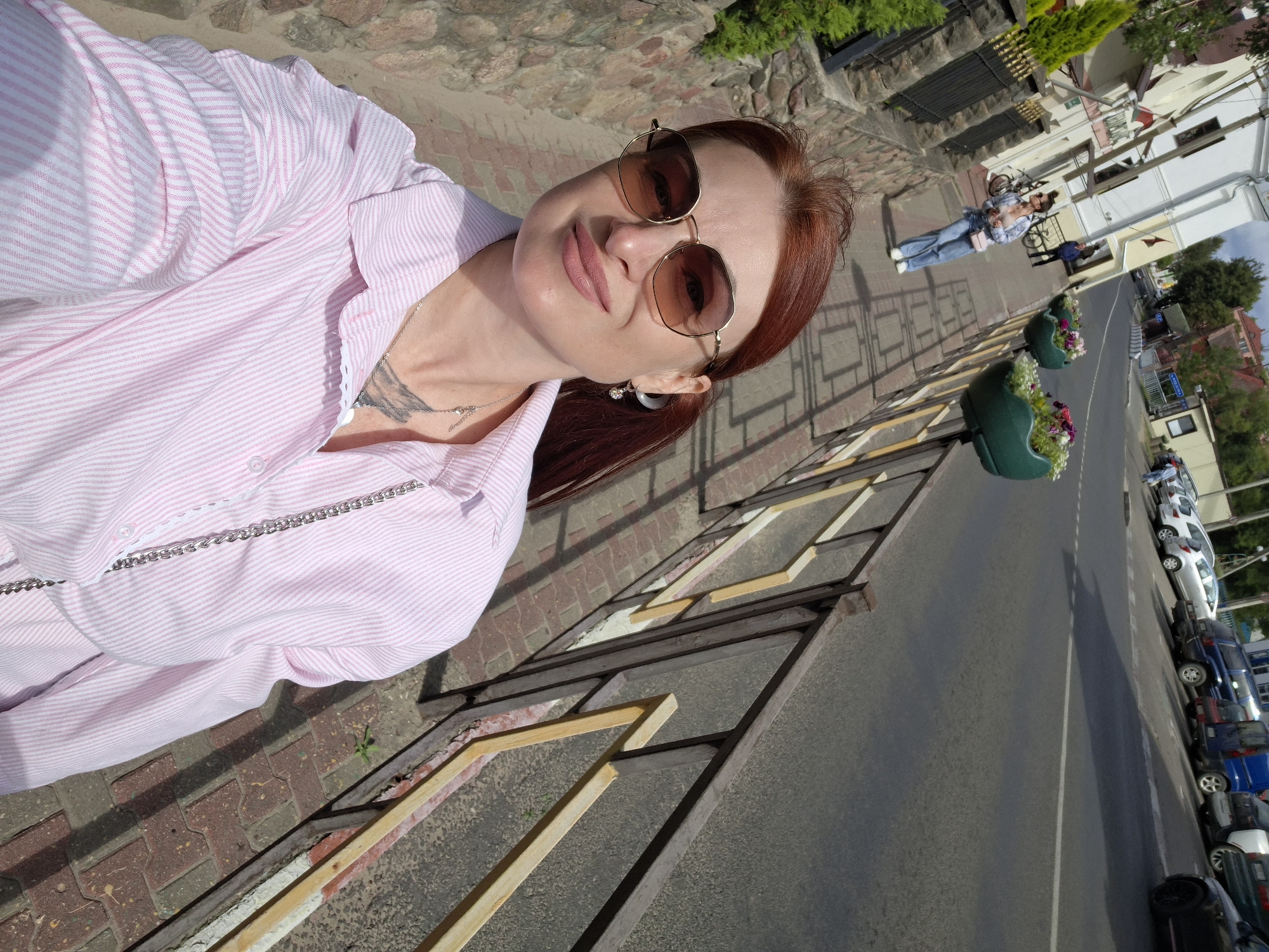 A woman with auburn hair, wearing a pink and white striped button-down shirt, sunglasses, and a necklace, takes a selfie while standing on a brick sidewalk. She has a small pink purse with a silver chain strap. In the background, a street lined with parked cars and a sidewalk with decorative planters is visible. The scene is set in Pastavy, Belarus.