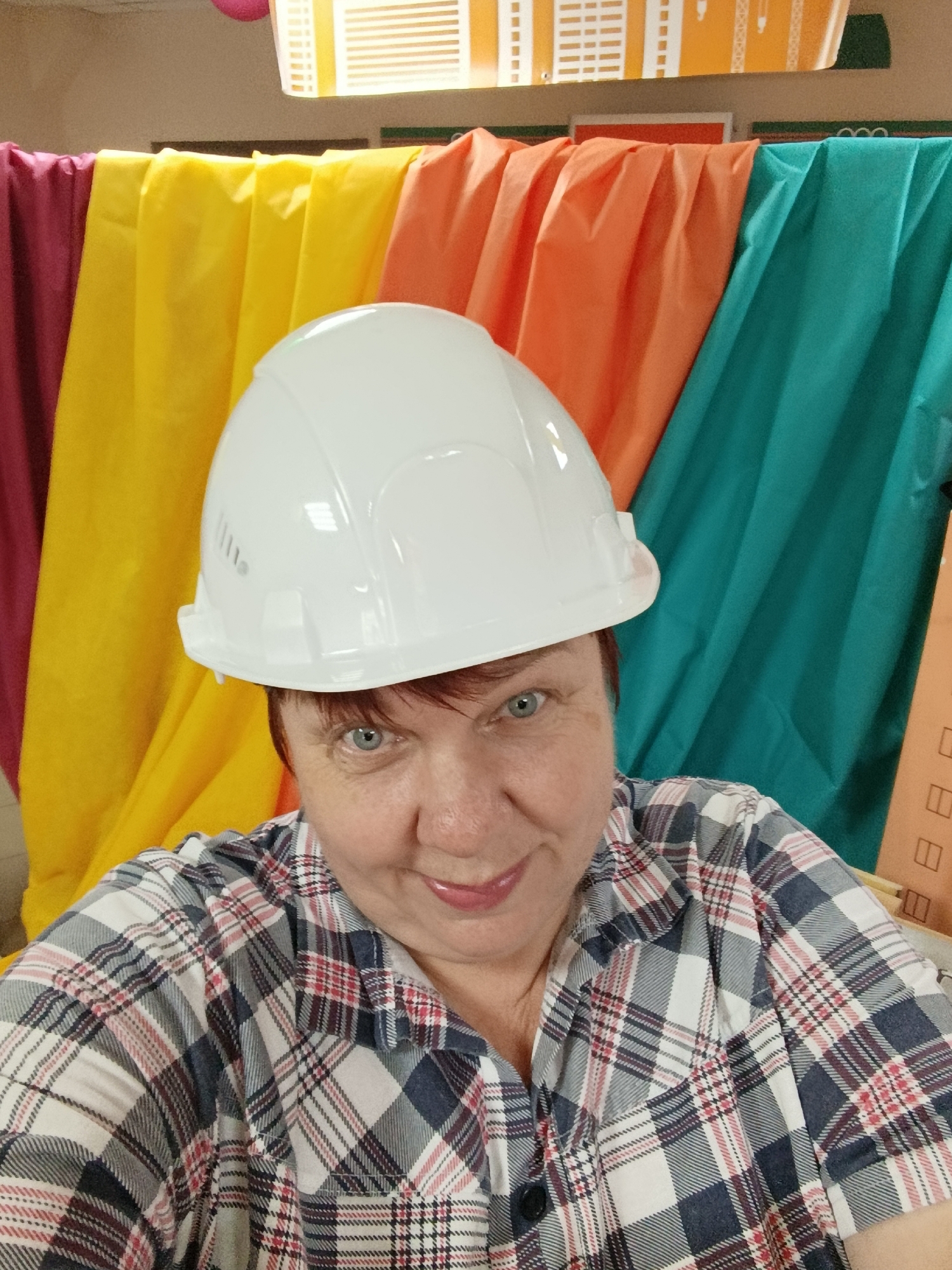 The image is a close-up, selfie-style photograph of a woman wearing a white hard hat and a plaid shirt. She has short red hair and blue eyes, and is smiling slightly. Behind her, there are brightly colored fabric backdrops in shades of purple, yellow, orange, and teal, draped in folds. The background appears to be indoors, possibly a decorated space like a party venue or a classroom. The woman's attire suggests she might be participating in an activity related to construction or a themed event. The location is identified as Svietlahorsk, Belarus.
