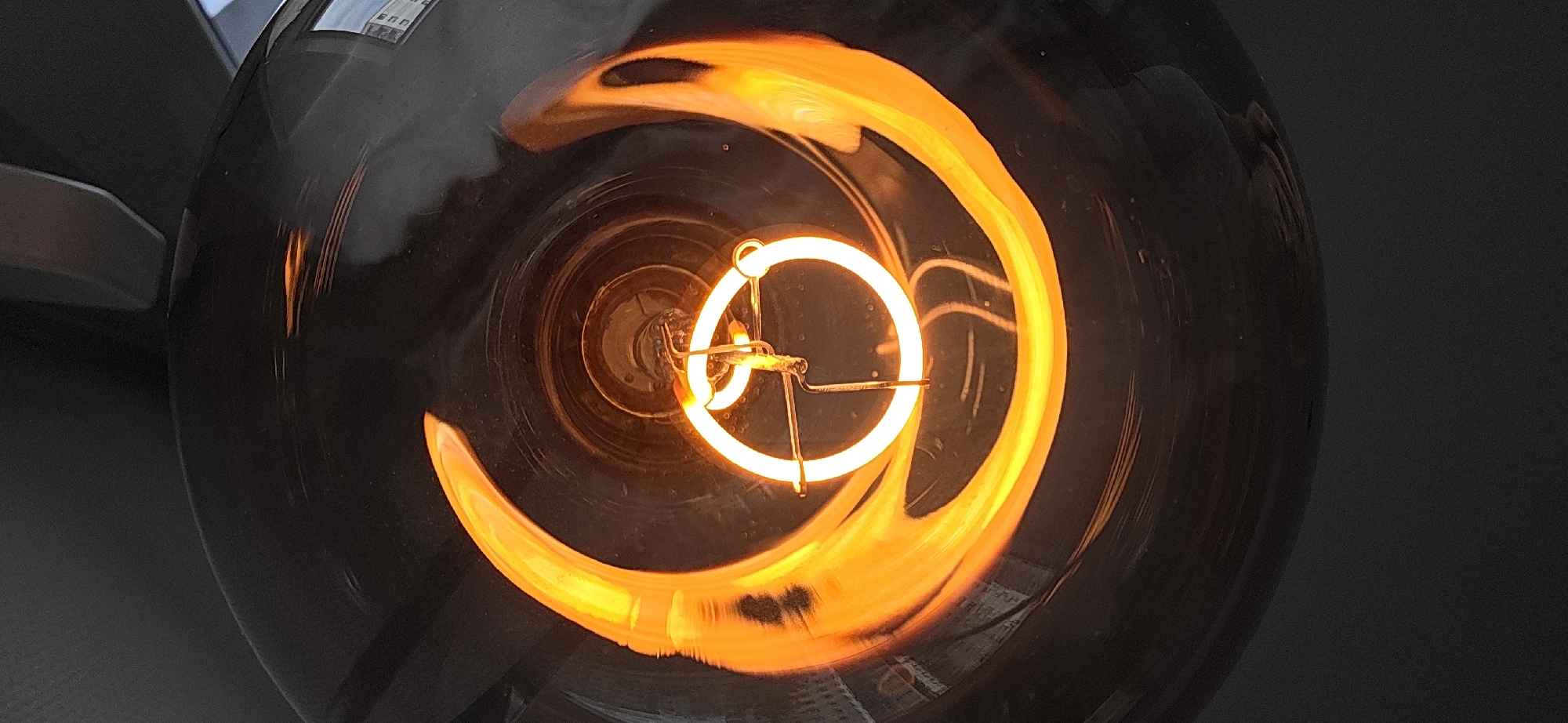 A close-up, overhead shot captures the filament of a lit Edison-style light bulb. The filament, a bright orange-yellow, forms a circular shape with a swirling pattern extending outwards. The filament is suspended by thin metal wires. The glass bulb surrounding it is dark, with reflections of the filament and surrounding environment visible on its surface. The background is dark and out of focus, suggesting the light bulb is the primary subject. The image does not provide any information about its location in Strasbourg, France.