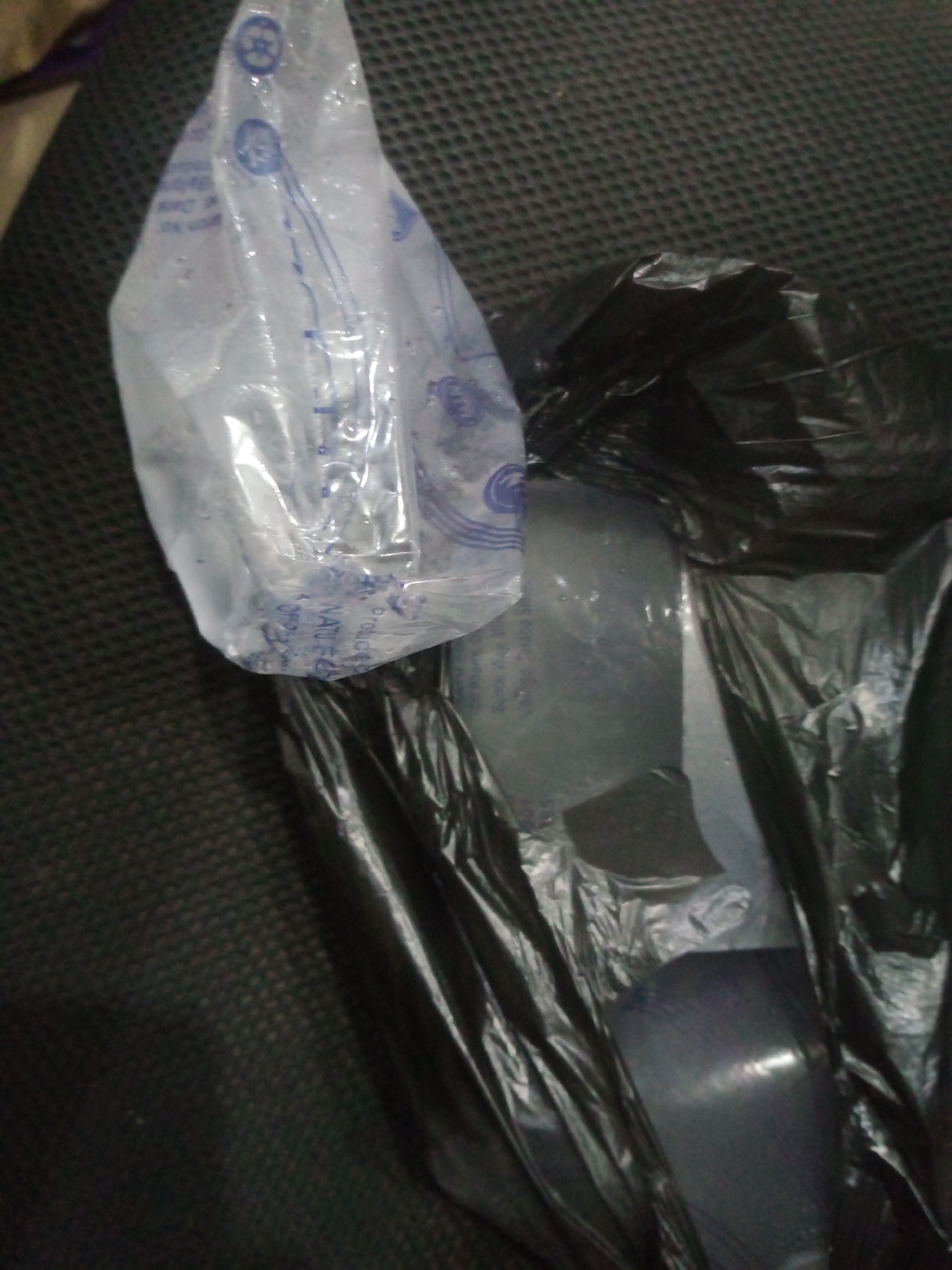 The image shows a close-up, overhead view of several items on a dark, textured surface. The primary objects are a clear plastic bag containing what appears to be ice or frozen water, and a black plastic trash bag. The clear bag is partially opened and is positioned on top of the black trash bag. The black trash bag is also partially opened, revealing more of the ice-like contents. Some of the text on the clear plastic bag is visible, but it is blurry and difficult to read. The overall lighting is dim, with some reflections visible on the plastic surfaces. The scene is indoors, likely a domestic setting, and there are no people visible. The location is Jalingo, Nigeria.