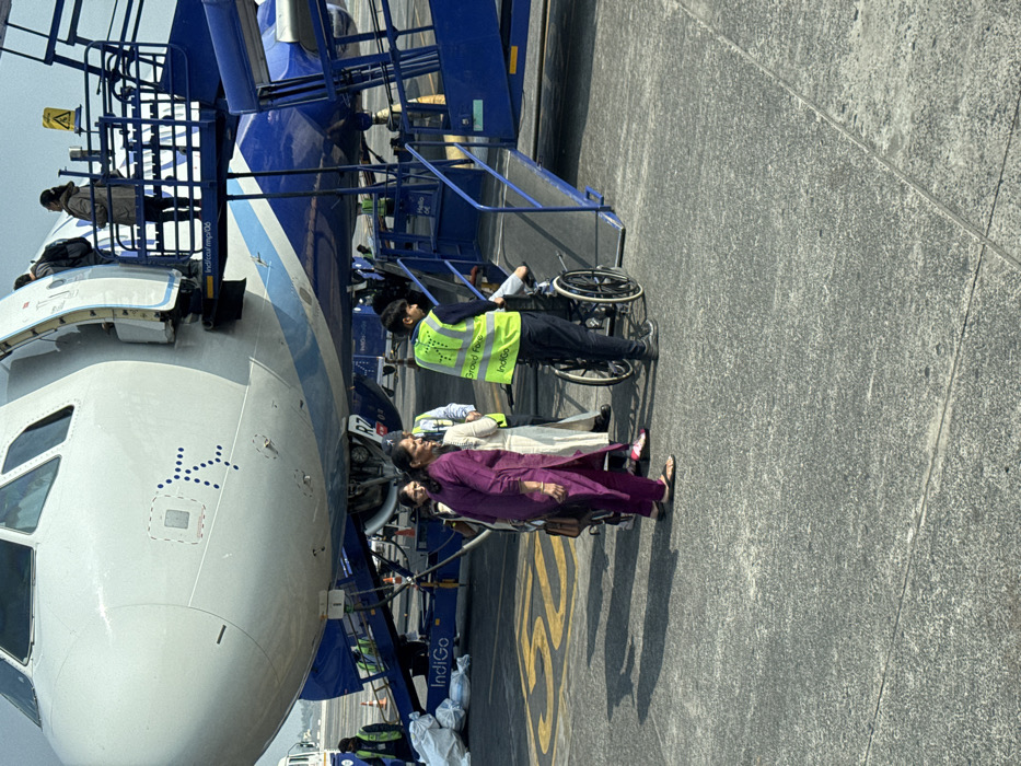 People are exiting an Indigo-branded airplane via stairs. A person in a wheelchair is being assisted by a ground crew member in a reflective vest. Two women in traditional Indian clothing are walking ahead of them. The nose of the plane is visible, with blue and white coloring. A yellow marking is visible on the ground.