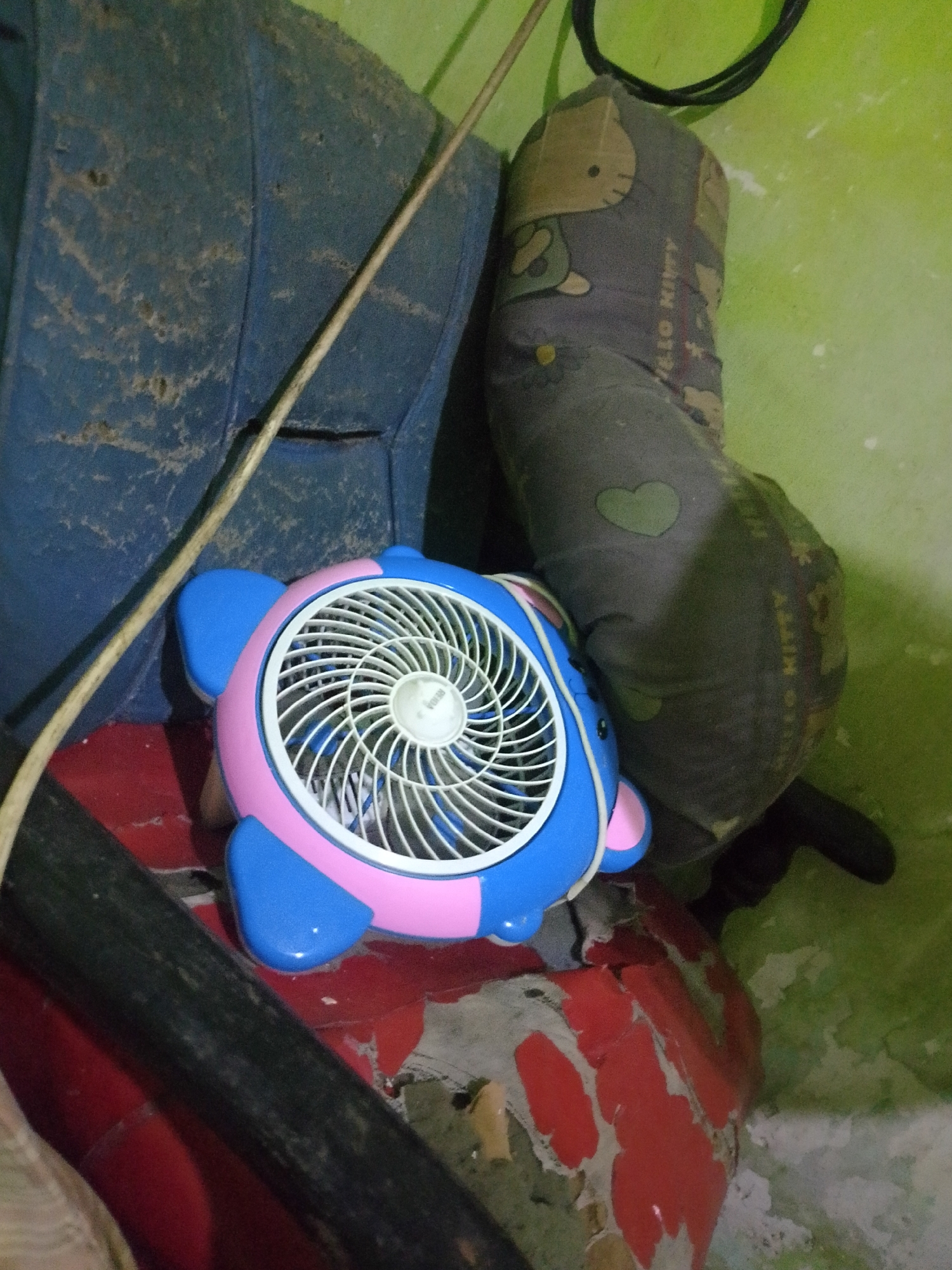 A pink and blue turtle-shaped fan is positioned on a red surface, likely a table or platform, within what appears to be a storage area or cluttered room. The fan is the central focus, with its white grille and blades visible. To the left of the fan, a large, dusty, dark blue object, possibly a cushion or bag, is leaning against a wall. Behind the fan and to the right, a grey, cylindrical object, which seems to be a pillow or bolster, is decorated with a pattern of Hello Kitty characters and a green heart. The wall behind these objects is painted a light green color and shows signs of wear and tear, with patches of white and grey exposed. A thin, light-colored cable runs diagonally across the image from the upper left towards the fan. The overall impression is one of a residential storage space in a tropical climate, given the presence of a fan and the general condition of the surroundings.