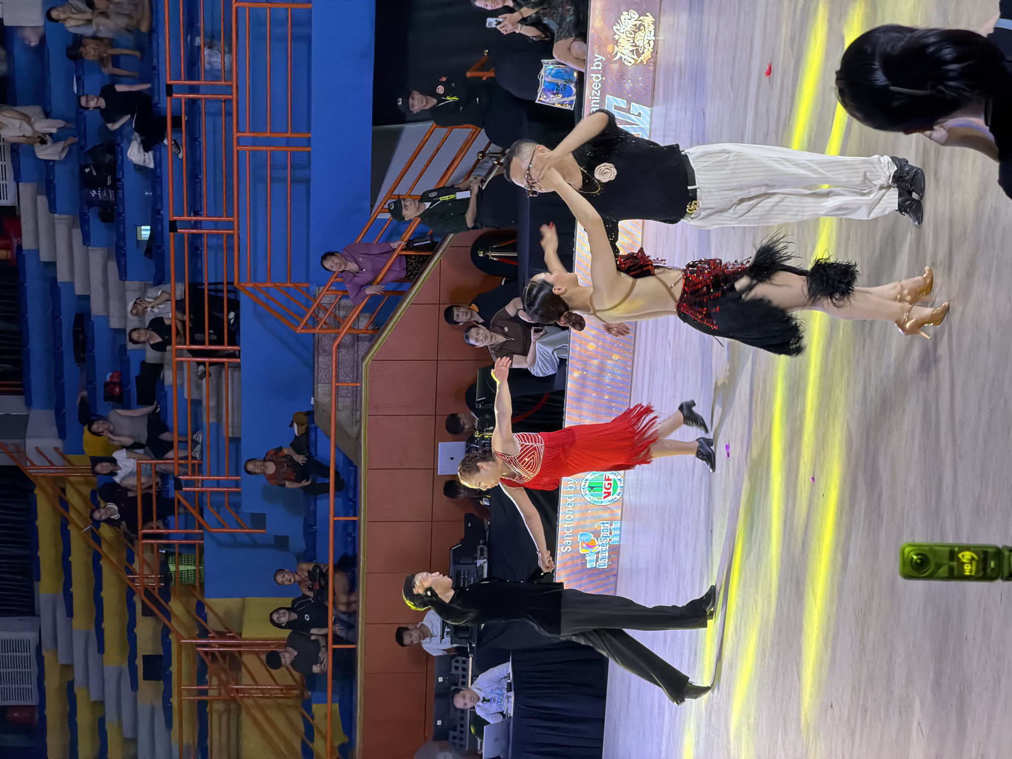 In this photograph taken in Hà Nội, Vietnam, a ballroom dance competition is underway. Four dancers, two couples, are the main subjects on the illuminated dance floor. The couple on the left features a man in a black suit and dark trousers, his arm raised, and a woman in a vibrant red fringed dress, her arms extended. To the right, another couple dances; the woman wears a dark, sequined dress with a feathered skirt, and the man is in a black sequined shirt and vertically striped trousers.
In the background, tiered seating is visible, occupied by spectators. Several individuals are seated in the blue and yellow bleachers. To the left of the dance floor, a table with laptops and equipment is manned by individuals, likely judges or officials. The event appears to be formally organized, with signage visible in the background that includes the logos of "Dancesport" and the Vietnam Dancesport Federation. A small, green HD camera is positioned on the floor in the foreground. The lighting on the dance floor consists of strong yellow beams. The overall environment suggests an indoor sporting or performance venue, likely an arena or stadium.