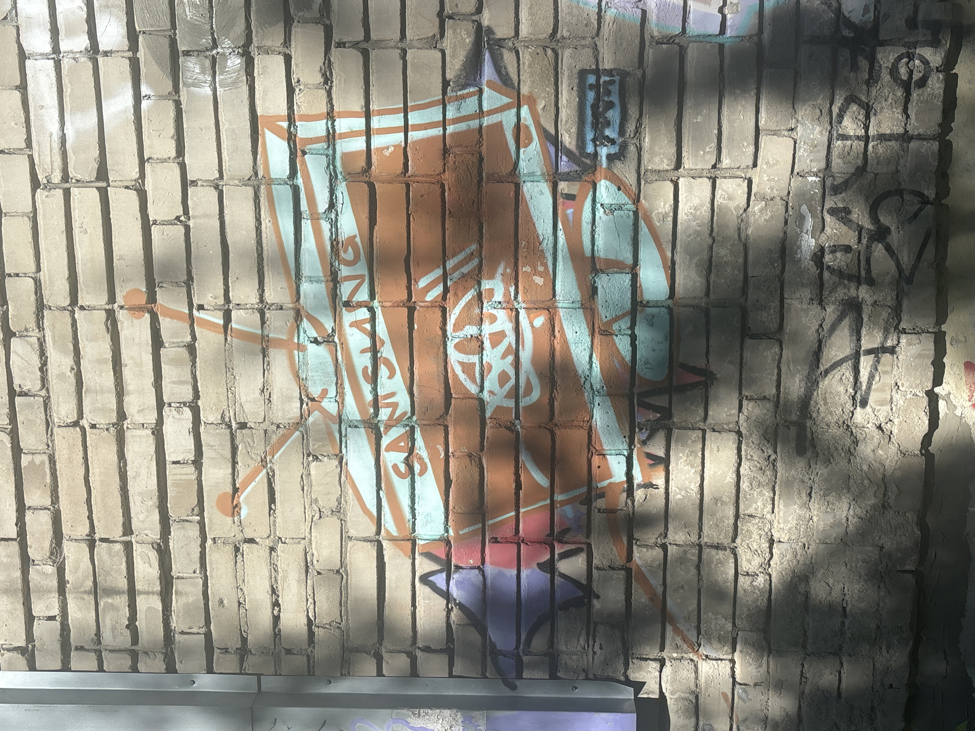 The image shows a close-up, eye-level, exterior shot of a brick wall with graffiti. The wall is made of light-colored bricks with visible mortar lines. The graffiti depicts a stylized television set with antennae, rendered in orange and light blue paint. The word "SAMSUNG" is visible in orange lettering across the screen of the television. There are also some abstract purple and pink shapes painted to the left of the television and some black graffiti below. Sunlight casts shadows across the wall, creating a dappled effect. The overall environment suggests an outdoor urban setting, possibly an alley or side street. The graffiti appears to be recent.