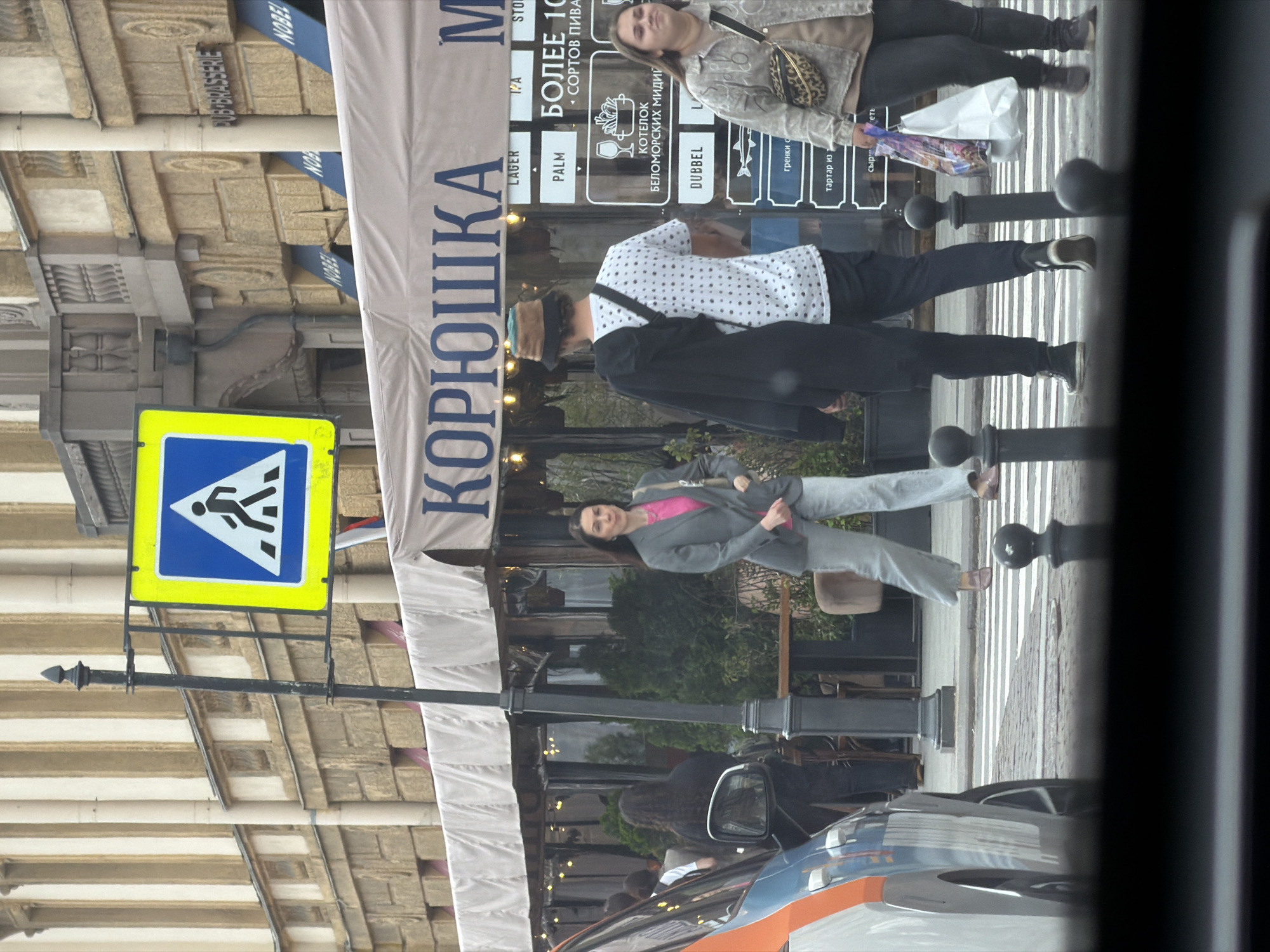 The image depicts a street scene in Lednevo, Russia, with a pedestrian crossing in the foreground. A blue and yellow pedestrian crossing sign is visible on a pole. In the background, a restaurant with a sign reading "КОРЮШКА" is present, with awnings and patrons visible through the windows. Several individuals are crossing the street. One woman in a gray suit and pink shirt is walking across the pedestrian crossing. A man wearing a patterned shirt, black pants, and a bucket hat is also present. Another woman, wearing a light-colored jacket with a leopard print fanny pack, is seen holding shopping bags. A car is partially visible on the left side of the frame. The building behind the restaurant has architectural details and signs, including "PUB-BRASSERIE" and "NOBEL".