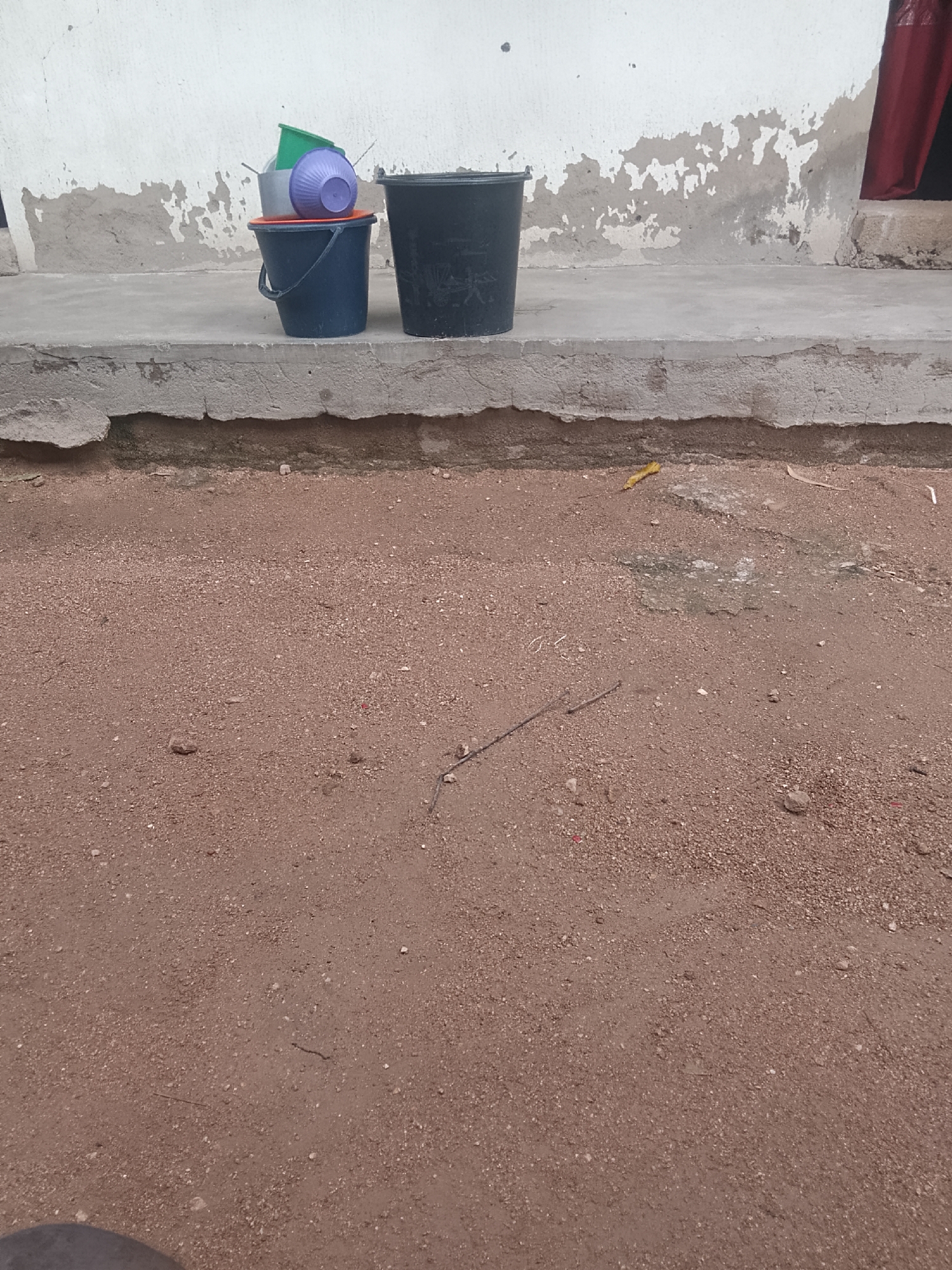 A close-up, eye-level shot shows a stack of plastic buckets on a concrete ledge in front of a weathered white wall. The buckets are blue, purple, and green, with one large black bucket to the right. The ground in front of the ledge is covered in reddish-brown dirt and small pebbles. The wall has peeling paint, and a red curtain is visible to the right of the black bucket.