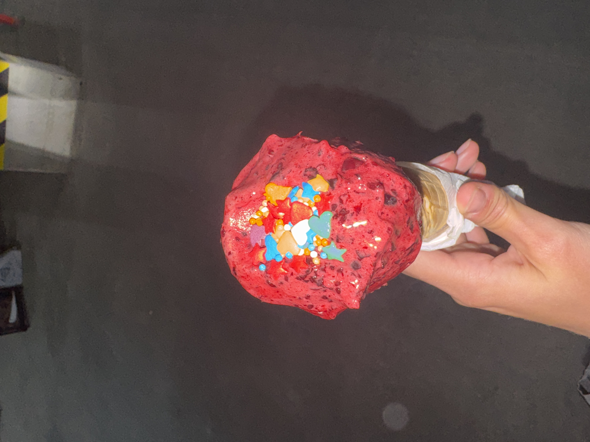 A hand is holding a very large ice cream cone, with a very large scoop of bright red ice cream that is heavily decorated with various colorful sprinkles and candies. The background is dark and appears to be asphalt, possibly a street or parking lot, in the city of Unknown, Unknown. In the top left corner, there is a structure with white and yellow markings on it.