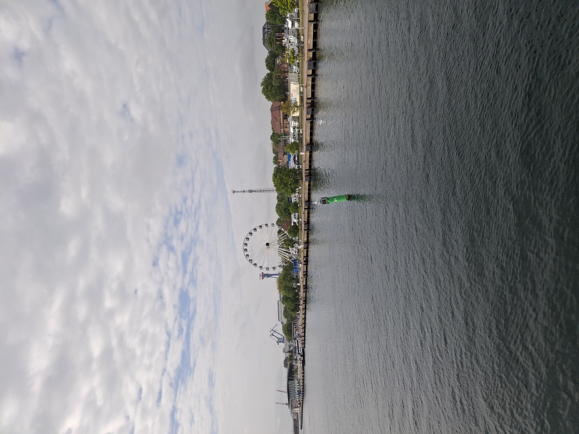 This eye-level shot shows a waterfront scene, likely in Rostock, Germany, given the presence of a Ferris wheel and industrial elements. The water occupies the right half of the frame, its surface reflecting the overcast sky above. The left half of the image features the shoreline, marked by a concrete barrier and a row of trees, behind which stand buildings. Notable landmarks include a large white Ferris wheel and various cranes. A green buoy is visible in the water near the shoreline. No people are readily discernible. The overall atmosphere suggests a commercial harbor or riverfront area on a cloudy day.