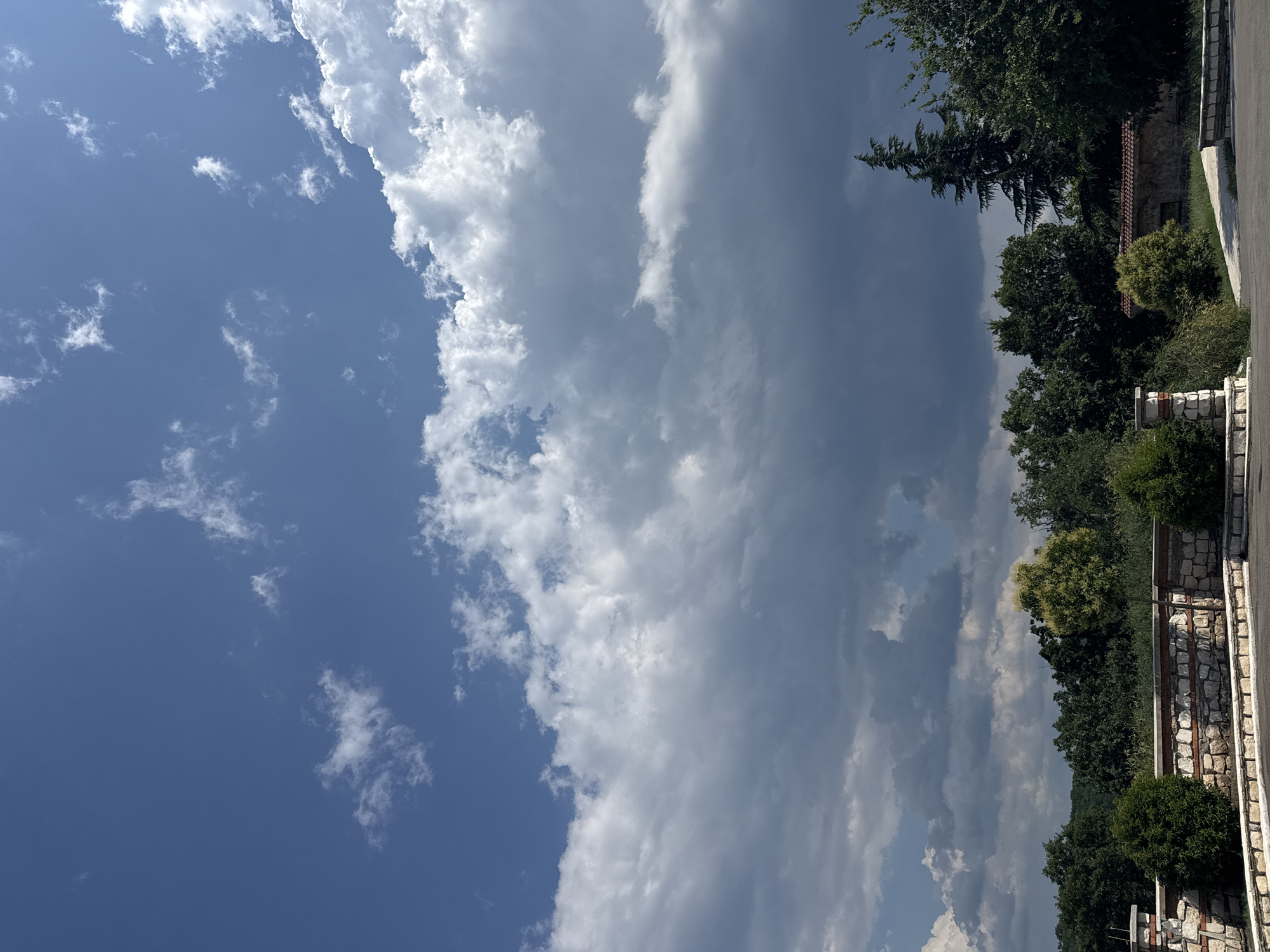 The image depicts a landscape with a dramatic sky over a rural or semi-rural setting. The sky is a prominent feature, with a mix of deep blue and heavy, dark grey clouds, suggesting an impending storm or the aftermath of one. White, fluffy clouds are also present, particularly in the upper portion of the sky, creating a contrast with the darker formations.
In the foreground, there is a stone wall, constructed from irregular light-colored stones with red brick accents. This wall appears to be part of a boundary or enclosure. Two small, manicured evergreen bushes are situated behind the wall, one on the left and one on the right, flanking a central section of the wall which includes a pillar-like structure also made of stone and brick.
Beyond the wall, a line of dense green trees forms a backdrop. The foliage varies in texture and shade, with some trees appearing taller and more mature, like the prominent conifer on the right side of the image. A small, stone building with a red-tiled roof is partially visible among the trees. A paved area, possibly a road or pathway, curves through the scene in the lower right. The overall impression is one of a natural landscape under a dynamic sky, possibly in or near Sredets, Bulgaria, given the provided context. There are no people or animals visible in the image.