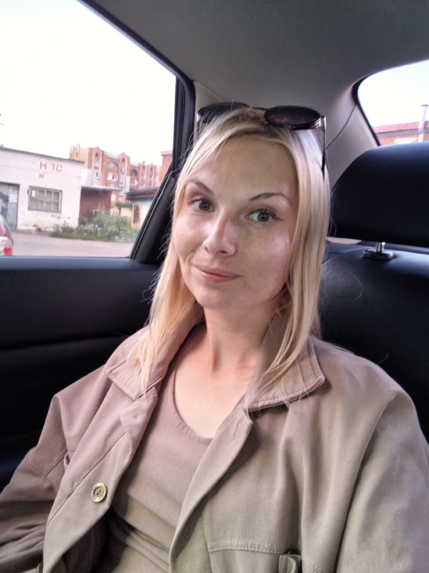 A blonde woman with light skin and freckles is sitting in the back seat of a car. She is wearing a light brown collared jacket over a beige top. Her sunglasses are perched on top of her head. The background shows a view out of the car window, revealing a street scene with buildings and some greenery. The buildings appear to be brick and some have industrial elements. One building has "H 10 H" written on it. The overall impression is a snapshot of a person in transit within an urban or semi-urban environment.