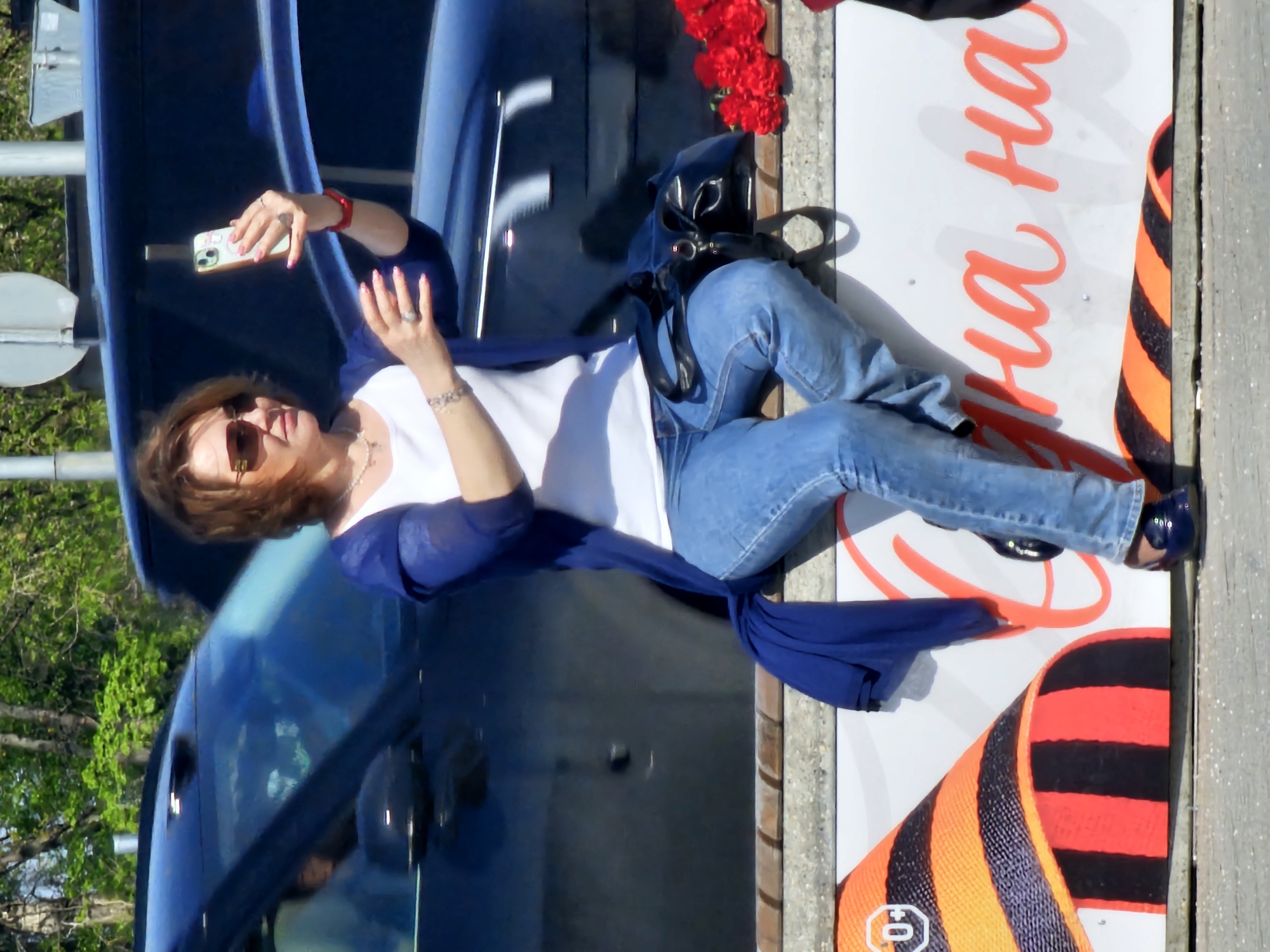 The photograph shows a woman seated on a sign adjacent to a car, seemingly taking a selfie. The woman, wearing sunglasses, a white top, blue jeans, and a blue cardigan, is holding a smartphone. A black purse sits near her. Next to her are red flowers. The sign she is sitting on features orange lettering and a black and orange ribbon pattern. The setting appears to be outdoors, with trees visible in the background and a glimpse of what looks like a street sign. Based on the presence of the ribbon and the Russian lettering, this may be a commemoration, likely in Moscow, Russia.