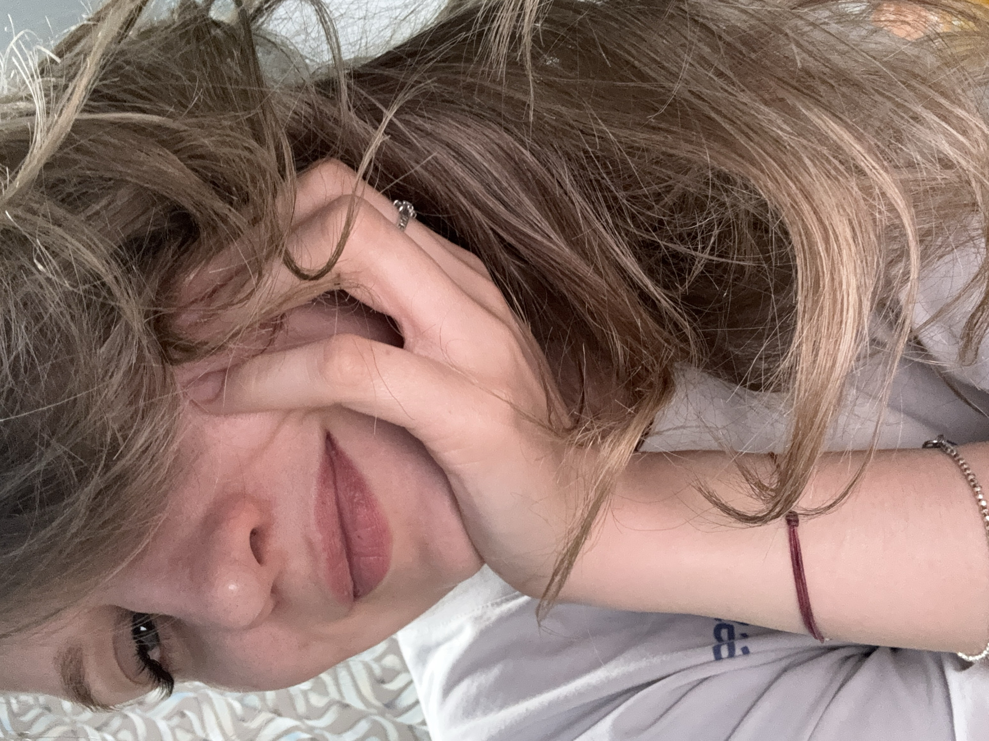 A person with light skin and light brown hair is lying down, partially covering their face with a hand. The person has a silver ring on one of their fingers. Two bracelets are visible on their left wrist, one red string and one silver. The person is wearing a light gray shirt. The background is a patterned surface. The image does not provide information regarding location in the city of Izhevsk, Russia.