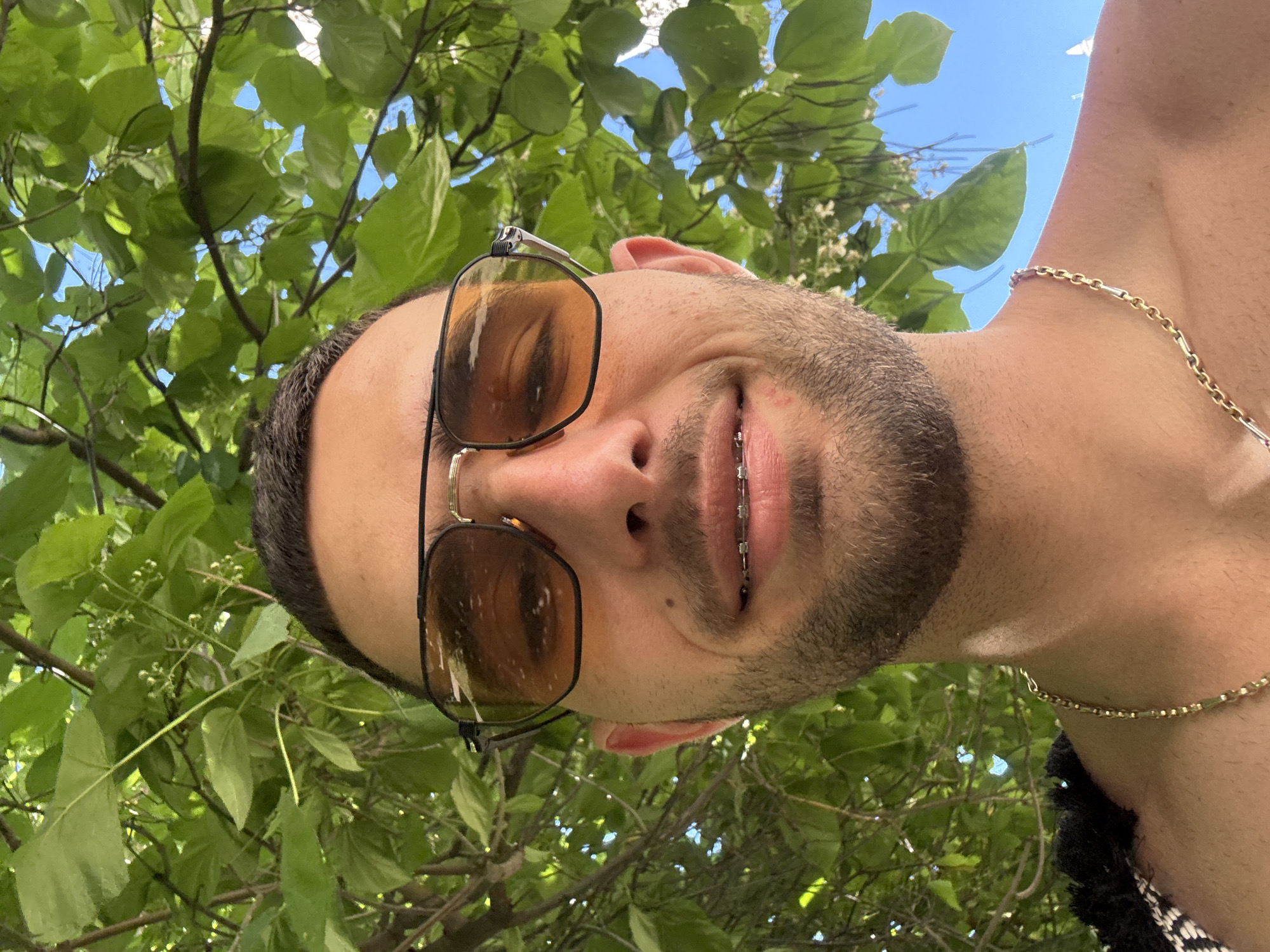 A close-up selfie captures a young man with fair skin and short dark hair, wearing orange-tinted aviator sunglasses. He has braces on his teeth and a light beard. He is shirtless, with a gold chain around his neck. The background is dominated by lush green foliage and a clear blue sky. The image appears to be taken outdoors, possibly in a park or garden, given the abundance of trees and leaves. The lighting suggests a sunny day. The scene is devoid of any specific urban or historical landmarks that would pinpoint it to Nesebar, Bulgaria, or any other specific city. The image is purely a portrait with a natural background.