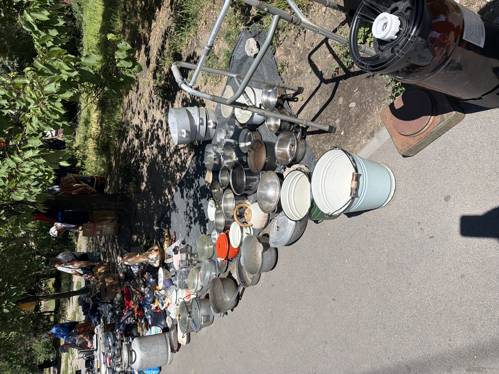 The image depicts an outdoor flea market scene, likely in Dnipro, Ukraine, given the context of similar markets in the region. A variety of used household items, primarily cookware, are displayed on a tarp spread on the ground and on the asphalt. The items include numerous pots, pans, buckets, colanders, and a large metal milk jug. Some people are visible in the background, appearing to be vendors or shoppers browsing the stalls. One man in a blue shirt is seated behind a display of goods. Another man in a white shirt and dark shorts is seated on a stool. A woman in a patterned headscarf and white top is standing. The market is set up under the shade of trees, with dappled sunlight filtering through the leaves onto the ground. A walker is positioned near some of the metal pots, suggesting potential accessibility needs or simply that it is also for sale. A large, dark plastic container with a white cap is also present. The ground is a mix of asphalt and dirt, with grass and vegetation visible in the background. The overall impression is one of a typical street-side market for second-hand goods.