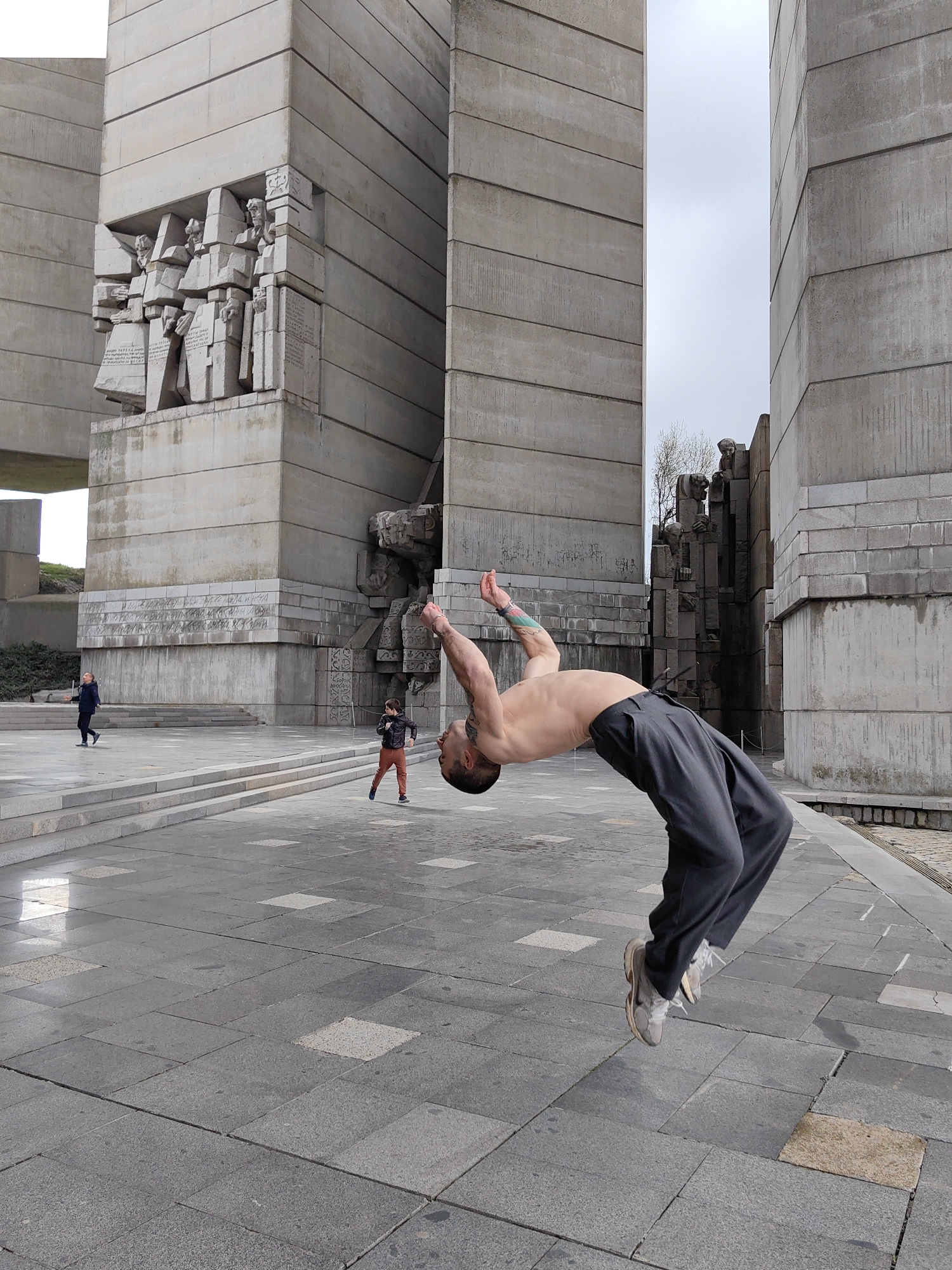 A man in gray pants and sneakers is performing a backflip in front of the Monument to the Founders of the Bulgarian State in Shumen, Bulgaria. The monument consists of large, gray concrete blocks with carved figures. In the background, a child in an orange jacket walks to the left, and another person walks in the distance. The ground is paved with gray stones. The sky is overcast.