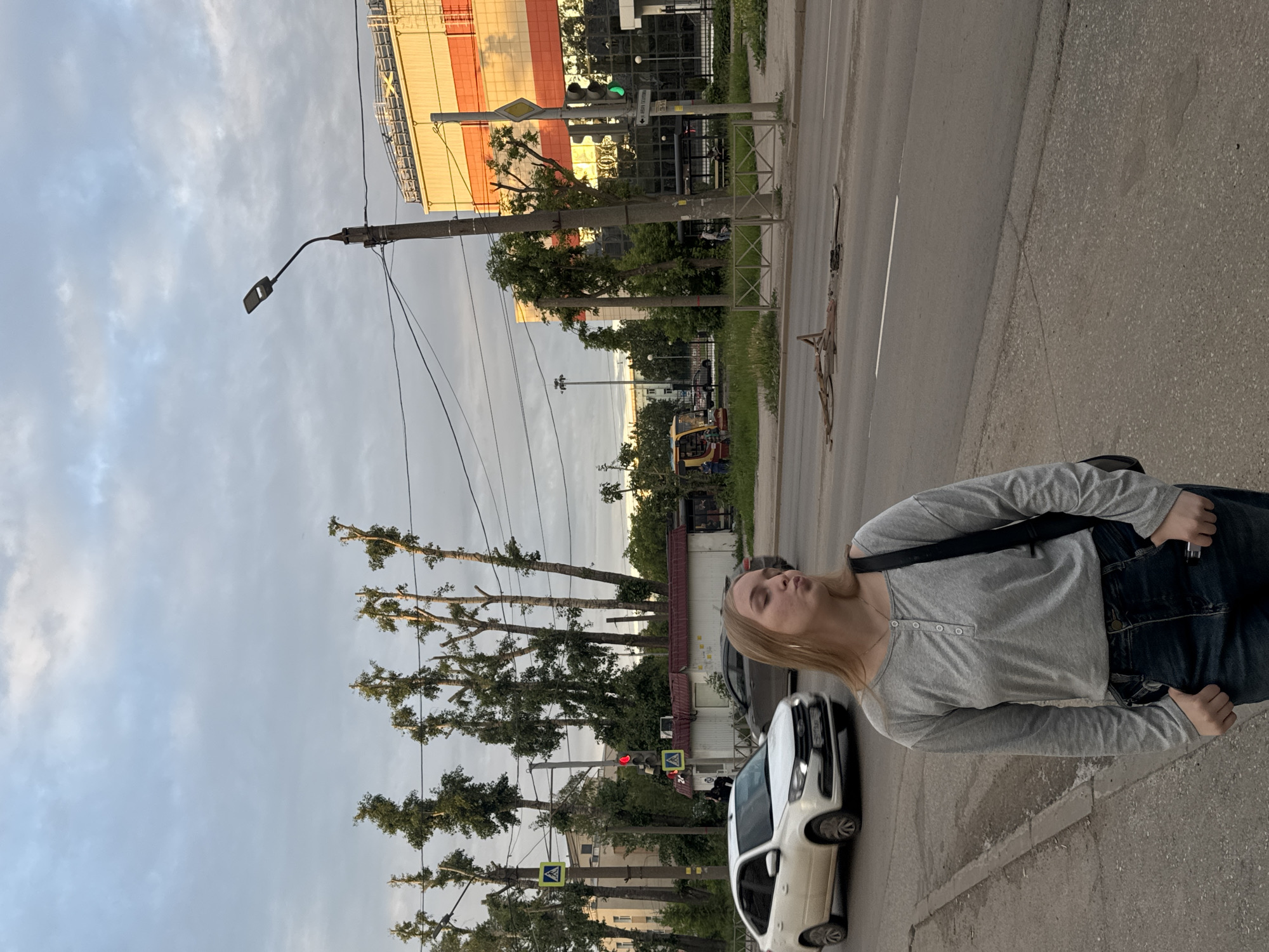 The image captures an urban street scene at the intersection of ulitsa Lenina and ulitsa Kuibysheva in Perm, Russia, likely during late afternoon or early evening under an overcast sky with patches of sunlight.
In the foreground, a Caucasian female with light brown hair stands on an asphalt sidewalk. She is wearing a grey long-sleeve Henley shirt with several buttons on the placket, dark blue jeans, and has a black bag slung over her left shoulder. Her hands are in her pockets, and she is making a puckering expression towards the camera. The sidewalk shows visible cracks.
To her left, on the multi-lane asphalt road, a white Lada Granta FL sedan with a Russian license plate (partially visible) is in motion, driving from left to right. A darker sedan is visible behind it.
Numerous tall, slender trees, many with their upper branches heavily trimmed or pollarded, line the streets. A grass median is present between road sections, on which a pile of broken wooden debris or planks is scattered.
Traffic infrastructure includes a red traffic light and a pedestrian crossing sign on the left side of the street, indicating a perpendicular road. On the right, a street sign beneath a green traffic light for the main thoroughfare clearly reads "ул. Ленина / ул. Куйбышева" (Lenin Street / Kuibyshev Street), confirming the location. A concrete utility pole with an attached streetlamp and overhead power lines is prominent on the right. Metal pedestrian barriers separate the sidewalk from the road.
Buildings visible include a small white kiosk with a red roof on the left side of the street near the intersection. On the right, the large, multi-story "Business Center Capital" building features reflective dark glass and distinctive alternating orange and white horizontal facade panels, illuminated by sunlight reflecting on its surface. In the background, a small yellow excavator suggests ongoing construction or maintenance activity. The sky above is largely cloudy with varying shades of grey.