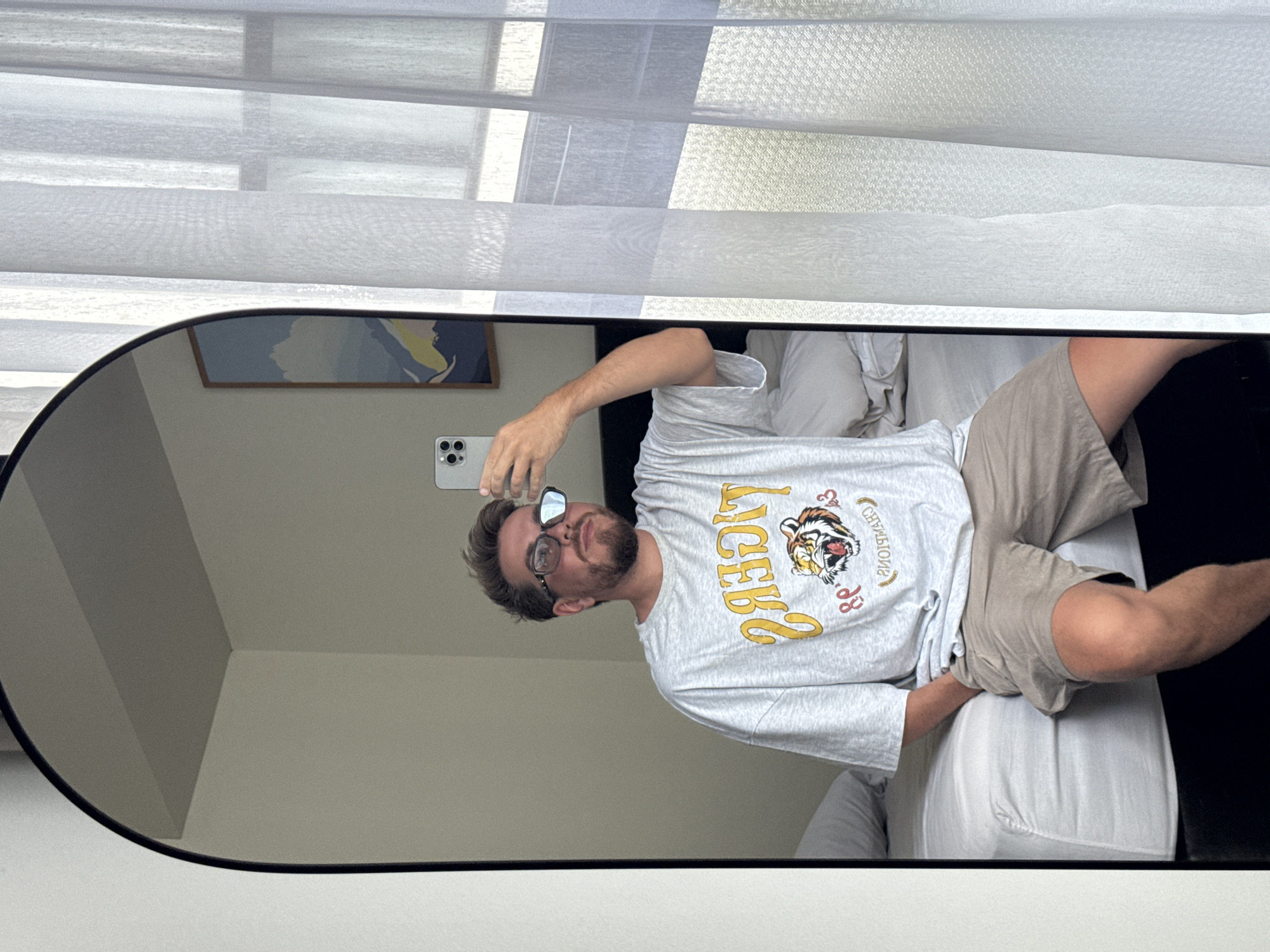 A man with a beard and glasses is visible in the reflection of an oval mirror. He is wearing a light grey t-shirt with a tiger logo and beige shorts. He is holding a smartphone in his left hand, presumably taking a selfie. The background shows a bed with white sheets and pillows. The mirror is positioned in front of a window covered with white blinds. A framed picture hangs on the wall in the background. The location is North Kuta, Indonesia.