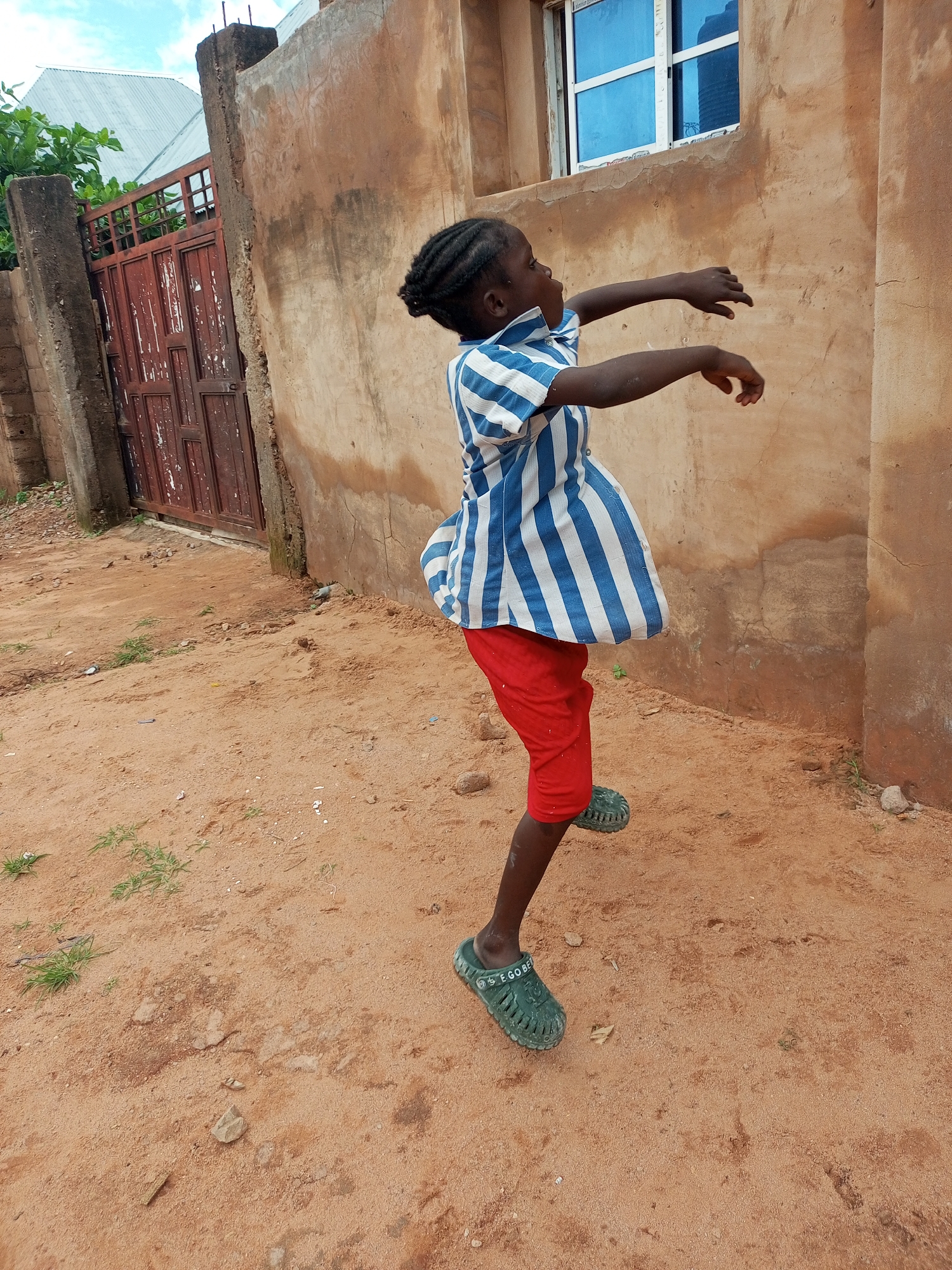 A young Black child, likely a girl given the dress, is captured mid-motion on a dirt path outside a building in Tempure, Nigeria. The child wears a blue and white striped shirt with a flared bottom and red shorts, and green clogs. Her arms are outstretched, and her body is twisted as if dancing or playing. The background features a weathered, ochre-colored building with a blue-paned window and a rustic, reddish metal gate with vertical slats. The ground is composed of loose, reddish-brown sand with scattered small rocks and sparse green vegetation. The sky above is overcast with a hint of blue. The scene appears to be in a residential or semi-urban area, suggested by the building and gate.