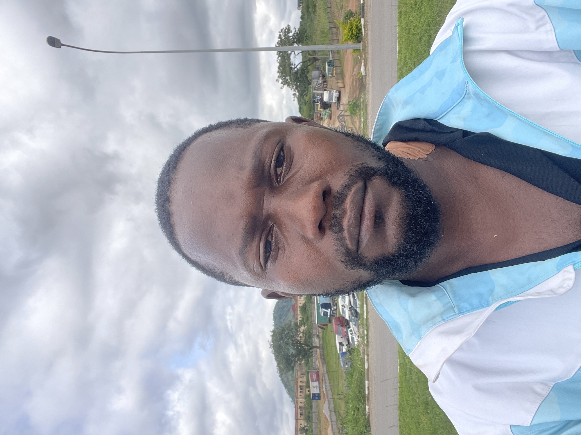 A medium shot photograph shows a Black man from the chest up. He is facing the camera directly, with his head slightly tilted. He has a beard and short hair. He is wearing a white and light blue jacket. The background shows a street, a grassy area, parked cars, buildings, and trees. The sky is cloudy. The location is identified as Kubwa, Nigeria.
