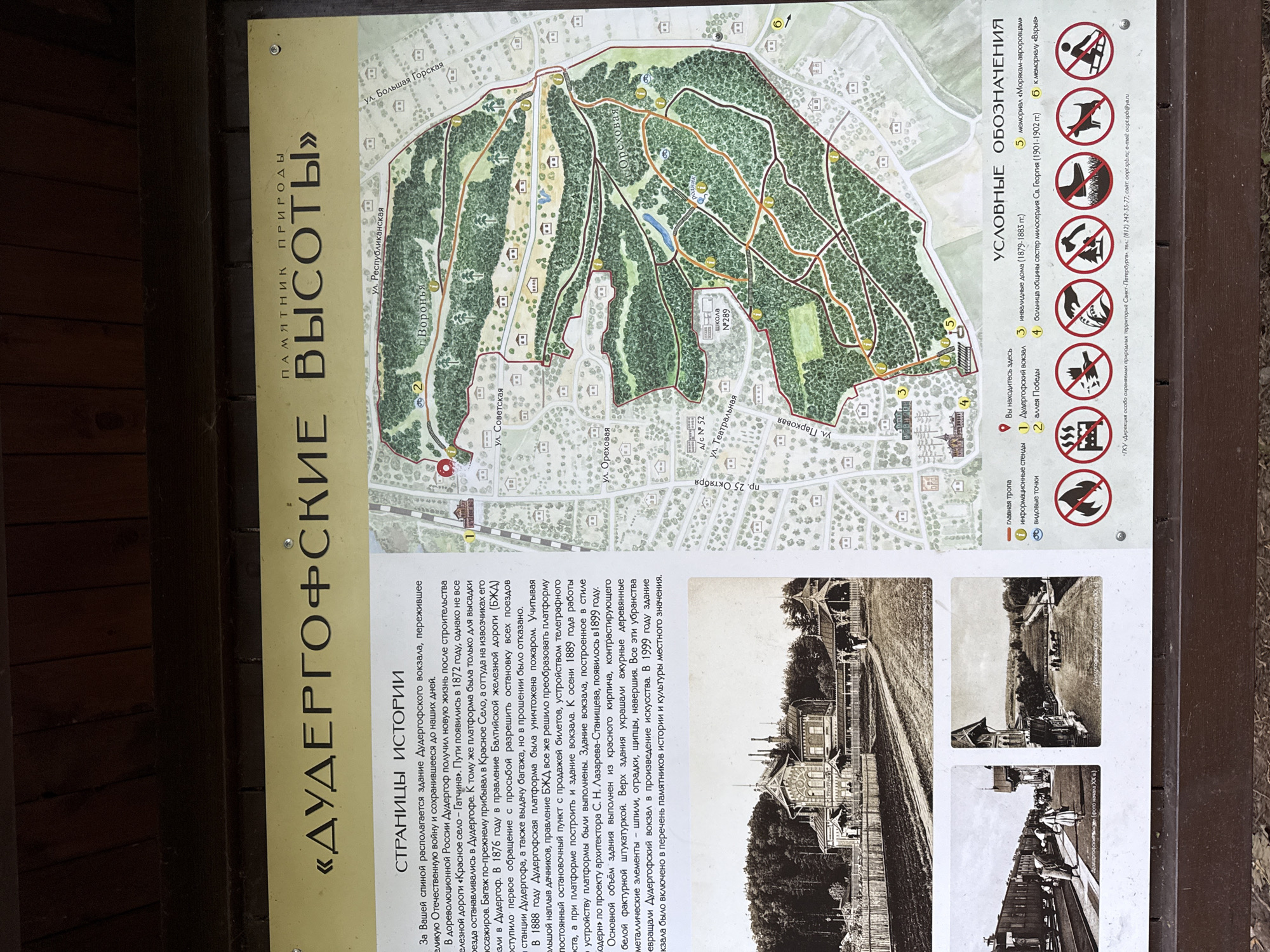 The image shows a historical informational display about a park in Krasnoye Selo, Russia. The display features a large, detailed map of the park with various trails and points of interest marked. Below the map, there are sections with historical text, old photographs of the area, and a legend of symbols used on the map.
The historical text describes the Duderhof station and its surroundings. It mentions the station's history, its role in the development of the railway, and its significance as a place of recreation for St. Petersburg residents. The text also touches upon the station's architecture and its preservation.
The old photographs depict the Duderhof station building in different periods, the railway platform with passengers, and a landscape view from the station.
The legend explains various symbols, including informational stands, the Duderhof station itself, Victory Alley, the St. George hospital, the "Moram-Avoruvam" memorial, and a memorial to "Explosion." There are also symbols indicating prohibited activities like fires and dogs off-leash, as well as a symbol for "You are here."
The environment depicted in the map is a park with wooded areas, paths, and some buildings. The overall impression is that of a historical site that has been preserved for visitors.