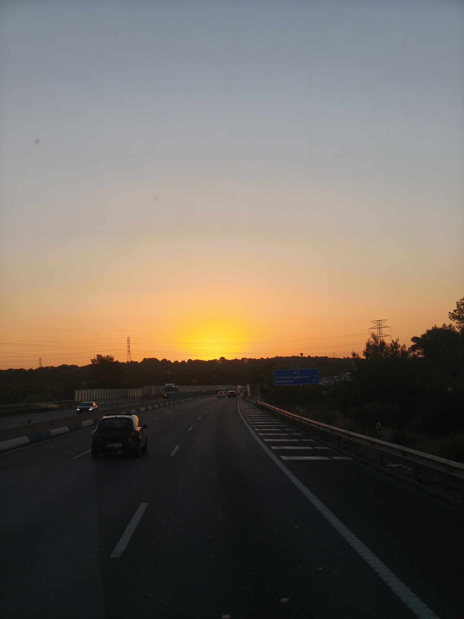 The image depicts a sunset viewed from a highway, likely the AP-7 or A-7, in the vicinity of Manises, Spain. The sun, a bright yellow orb, is low on the horizon, casting a warm orange and yellow glow across the sky. The sky transitions from a pale blue at the top to a deep orange near the sun.
The highway itself is a multi-lane asphalt road with clear white lane markings. Several cars are visible on the road, driving in the same direction as the camera. The closest vehicle in the foreground is a dark-colored hatchback. Guardrails line the side of the highway, and on the right side, a dense line of trees and foliage is visible, with tall electrical pylons interspersed in the background.
A blue highway sign is visible on the right side of the road, indicating directions to "Castelló/Castellón" and "Barcelona." The surrounding landscape appears to be somewhat rural, with a gentle, hilly terrain visible behind the trees. The overall atmosphere is serene, with the warm light of the sunset creating a tranquil scene on the highway. There are no people clearly visible in the image, and the focus is on the landscape and the highway.