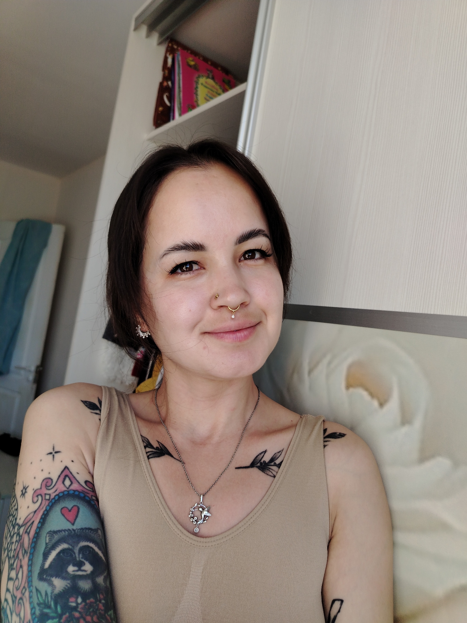 A woman with dark hair is positioned in the foreground, exhibiting a slight smile. She is wearing a beige tank top and accessorized with earrings, a nose ring, and a necklace. The tattoos on her arms and upper chest are visible. In the background, the elements suggest an interior setting. There are cabinet doors and shelves, and what appears to be an indistinct floral design, possibly a picture. There is no definitive information in the image to indicate that it was taken in the city of Noviy, Russia.