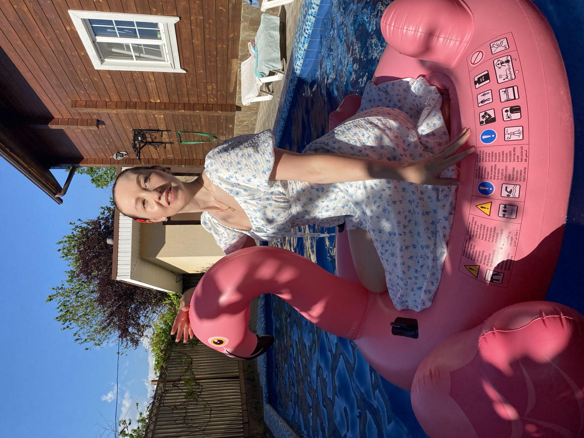 This image captures an outdoor residential scene in Dagomys, Russia, under clear, sunny skies, likely during late morning or early afternoon given the high sun angle and sharp shadows.
In the foreground, a female individual, appearing to be an adult in her 20s to 30s, is seated on a large, pink inflatable flamingo in a blue-tiled swimming pool. She has fair skin, light brown hair secured with a red clip on her right side, and is wearing a white, short-sleeved, square-necked dress with a blue floral pattern. A thin gold necklace is visible around her neck. Her left hand rests on the flamingo's neck, while her right hand is placed on its body. She is looking slightly to the viewer's left with a subtle smile.
The inflatable flamingo, branded "INTEX" on its neck, features yellow eyes with black pupils and a black beak. Its body contains multiple black-on-yellow and black-on-white warning pictograms and multilingual text (including English, German, French, Italian, Spanish, Russian, and Arabic), indicating safety information such as "inflatable swimming aid," "not for swimmers," "max 40 kg," and "adult supervision required." A black valve is present on its top surface.
The swimming pool features light blue and dark blue mosaic tiling around its edge and is filled with clear, rippling water reflecting the bright sunlight.
In the background, to the right, stands a two-story residential building constructed of dark brown horizontal wooden paneling. It has a white-framed window with multiple panes that reflect the sky, and a gutter system along its roofline. A black wall-mounted pull-up bar with two green straps is affixed to the wooden wall. To the left, behind the pool, is a lighter-colored, possibly stucco or paneled structure with a white overhang/soffit. Further to the left, a light brown wooden slatted fence is visible, partially obscured by green vegetation and some vines. Various trees and bushes, including one with both green and reddish-brown foliage, populate the background under a clear blue sky dotted with a few wispy white clouds. On the paved area beside the pool to the right, two white plastic lounge chairs are visible, one covered with a blue towel and another item.