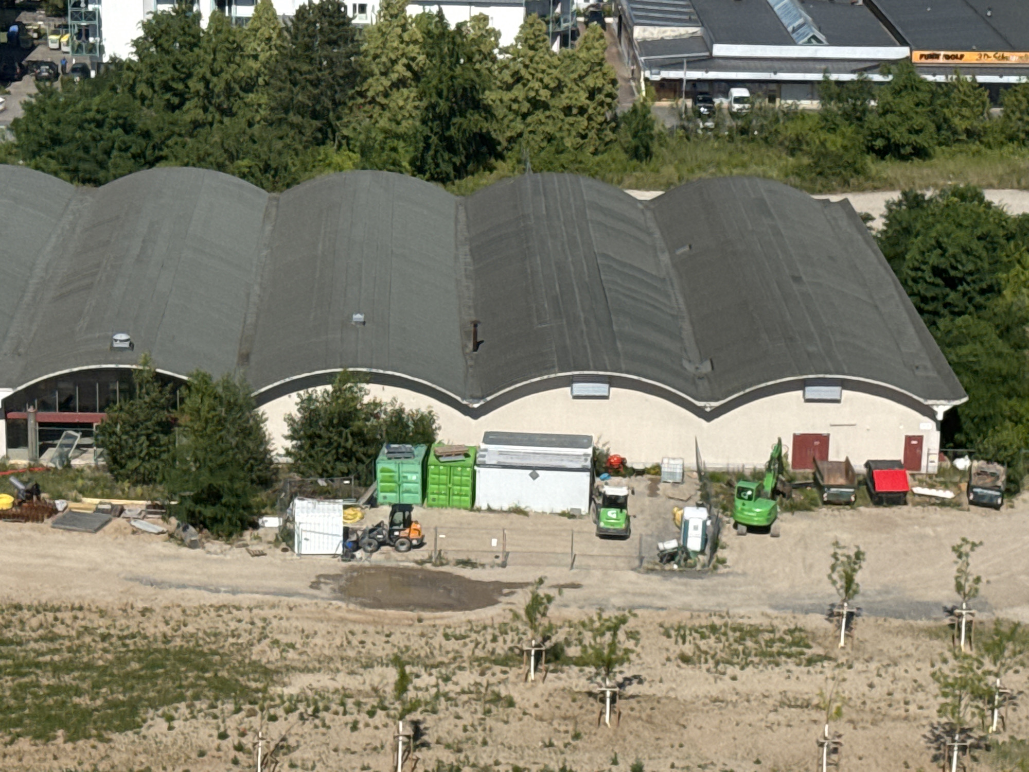 This is an aerial, slightly angled, wide shot of a construction or industrial area in Mannheim, Germany, with a focus on a large, corrugated, arched-roof building. The building has a dark grey roof with visible seams and several skylights. The facade is light-colored, with arched windows or openings at the top and red doors on the right side. In front of the building, there is a gravel and dirt lot with various construction vehicles and equipment. To the left, there is an orange mini-excavator next to a collection of green shipping containers. In the center and right of the lot, there are more construction vehicles, including a green excavator, a green dump truck, a red-topped dumpster, and a dark colored vehicle. Some portable toilets and storage units are also visible. The area is surrounded by trees and vegetation, with glimpses of other buildings and a road in the background. Several newly planted saplings with protective stakes are present in the foreground, suggesting ongoing landscaping or site development.