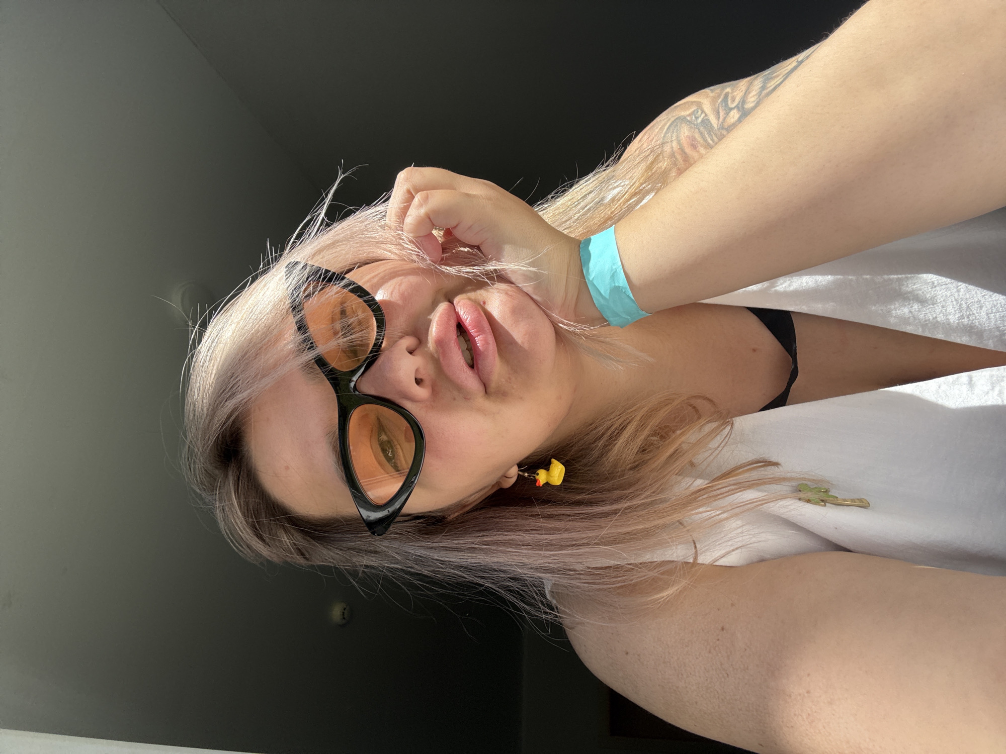 A medium shot captures a woman with light pink hair and large, dark-rimmed, orange-tinted cat-eye sunglasses. She is resting her chin on her left hand, with her fingers brushing her cheek. She wears a white tank top over a black bra, and a turquoise wristband is visible on her left wrist. A small yellow rubber duck earring dangles from her left ear. Her mouth is slightly open. The background is dark, suggesting an indoor setting with light coming from the right, casting shadows. The image does not provide any specific visual information that would allow for its placement within the city of Birobidzhan, Russia.