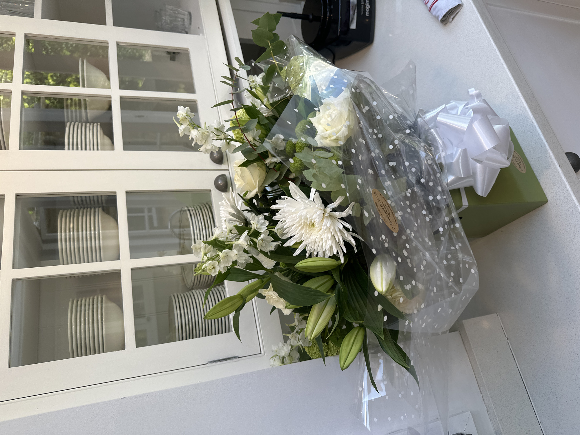 The image shows a bouquet of white flowers wrapped in polka-dotted cellophane with a white bow. The bouquet is placed on a white surface, possibly a countertop. In the background, there is a white cabinet with glass doors, revealing stacks of white plates with black stripes. A black appliance is partially visible above the bouquet. The location details "Unknown, Unknown" do not provide specific information about the city or setting.