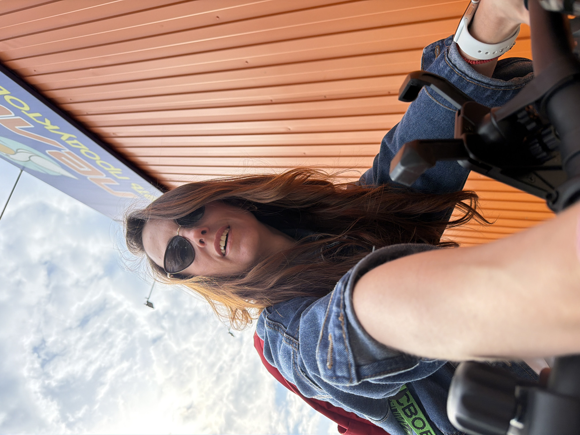 A woman with long brown hair and sunglasses is taking a selfie. She is wearing a denim jacket over a red top, a white wristwatch, and a red bracelet. She is holding a black object, possibly a camera or phone accessory. The background shows a cloudy sky and the edge of a building or structure with orange paneling. To the left, there is a sign with Cyrillic lettering, indicating the location is likely in Russia. Based on the Cyrillic writing, the location is likely in the city of Birobidzhan, Russia.