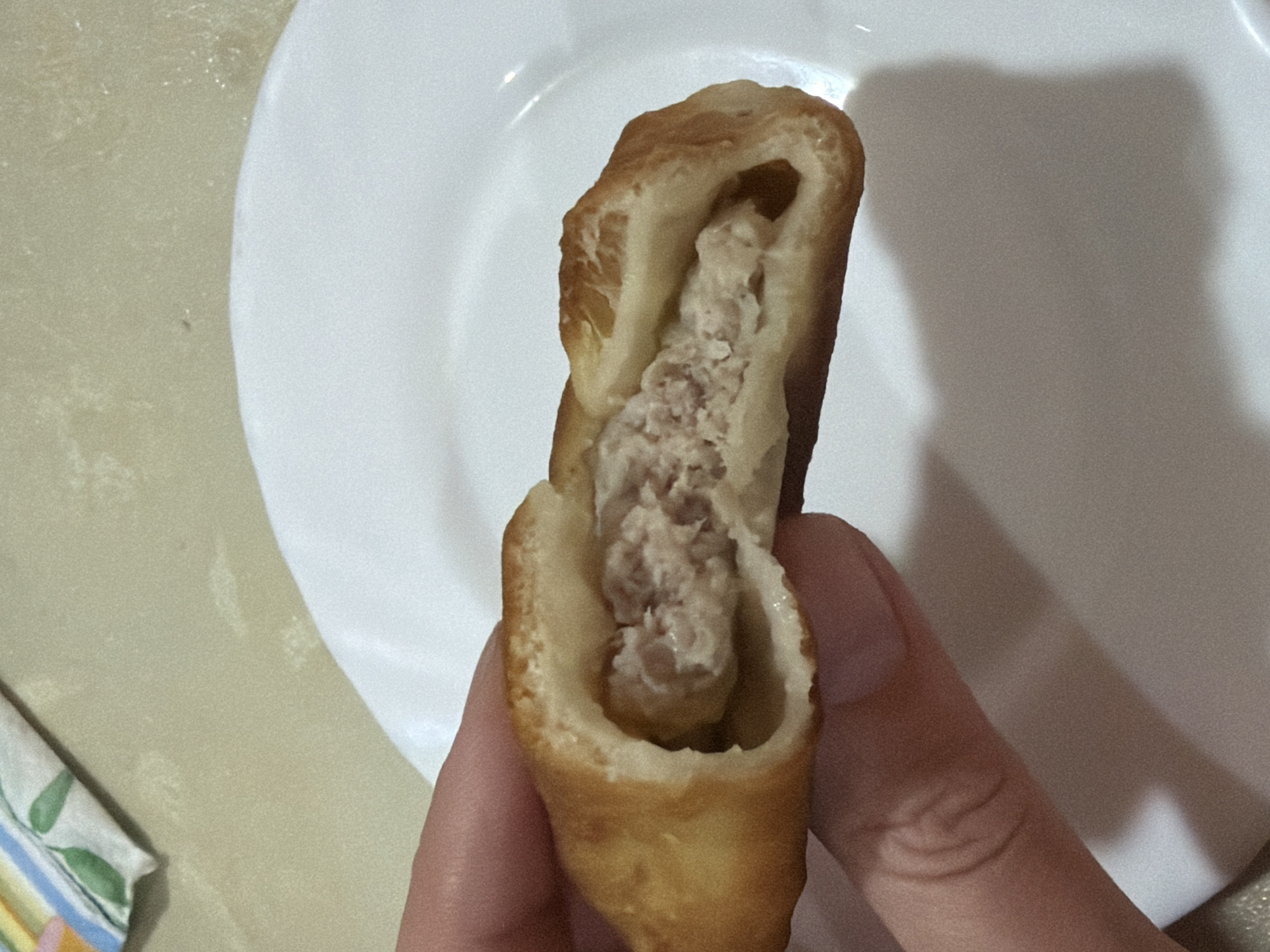 A hand holds a fried pastry filled with meat, which is broken open, revealing the filling. A white plate is partially visible behind the pastry. A patterned napkin lies on a surface to the left of the plate. The location is Chaikovsky, Russia.