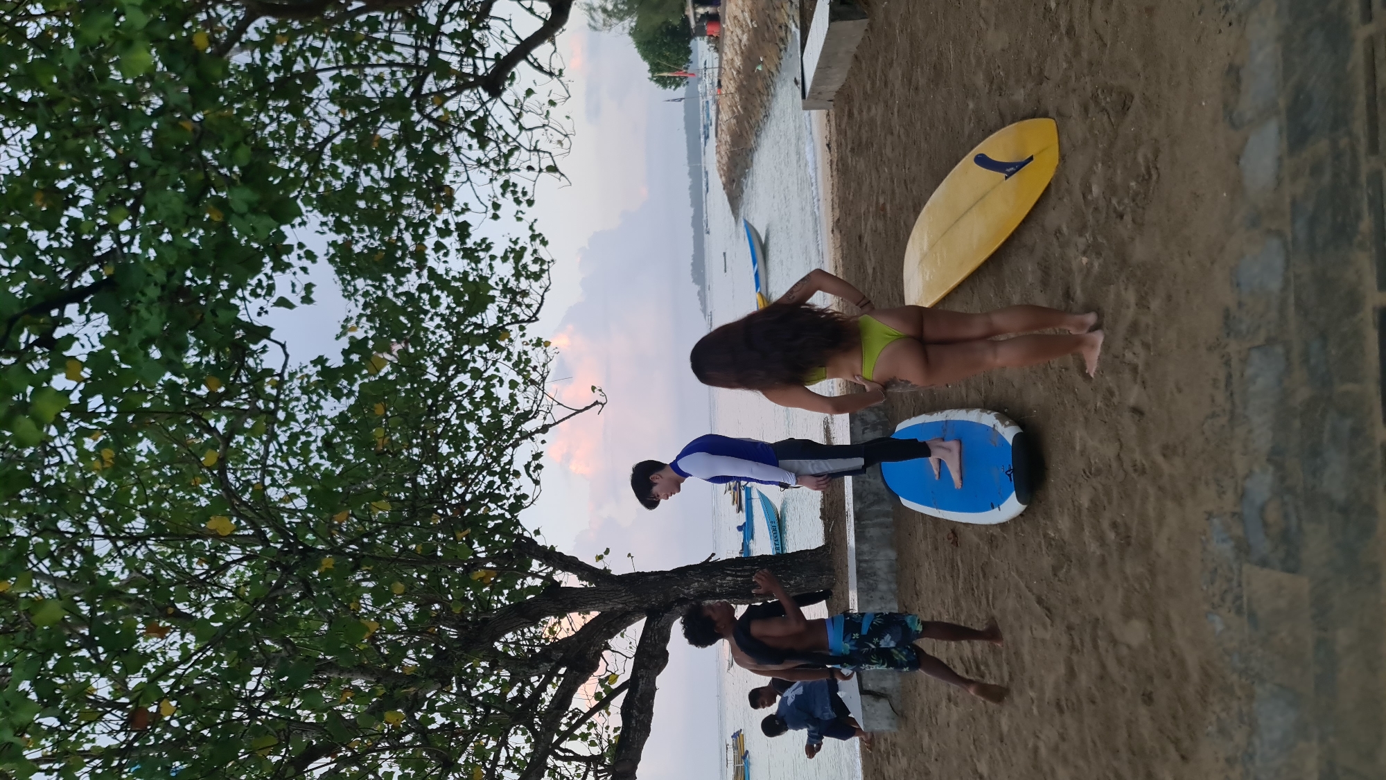 A group of young people are gathered on a sandy beach in Denpasar, Indonesia, near the water. The sky is overcast, with hints of pink and orange in the clouds, suggesting either sunrise or sunset. In the foreground, a man in black swim shorts with a blue and white pattern stands facing a person in a blue and white long-sleeved rash guard and black leggings. The person in the rash guard is standing on a blue surfboard. To their right, a woman in a neon green bikini faces away from the camera, her back to the other two. Next to her lies a yellow surfboard. In the background, several boats are anchored in the calm water, and there is vegetation on the shoreline. To the left, two people are seated on a low concrete wall. The overall atmosphere appears relaxed and recreational, with the surfers and their equipment indicating a water-sport activity.
