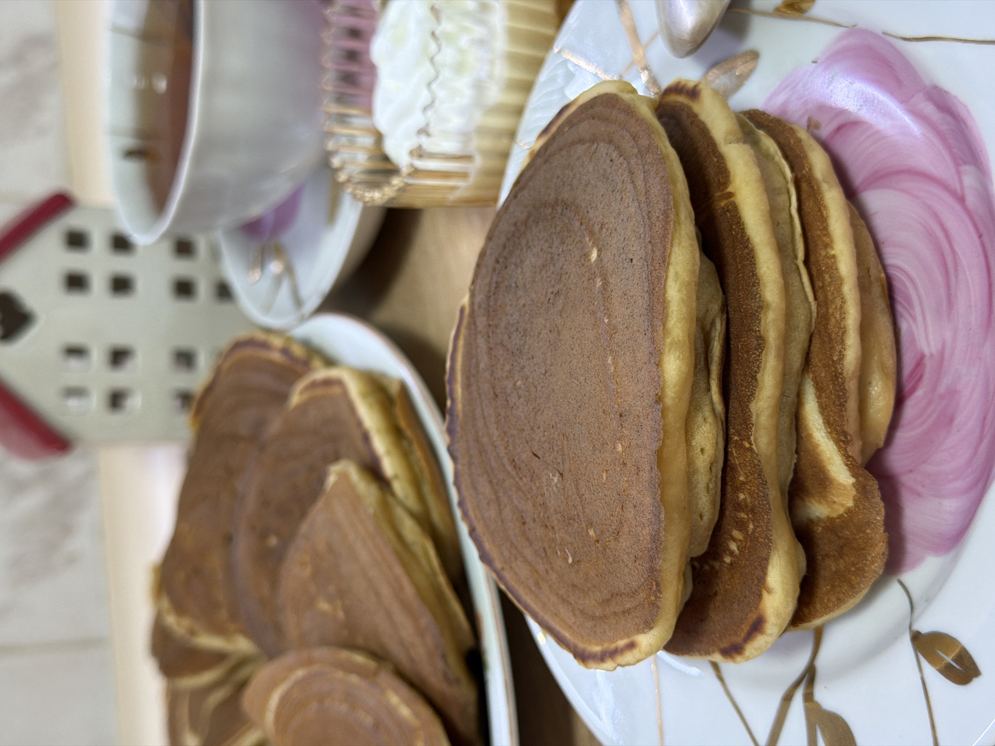 A close-up shot captures a stack of golden-brown pancakes on a white plate adorned with pink floral designs. The pancakes are slightly uneven in shape, with visible texture on their surfaces. To the right of the pancakes, a small glass bowl contains a dollop of white cream or yogurt. In the background, a white teacup with a delicate pink pattern rests on a saucer, filled with amber-colored tea. Behind the teacup, a miniature white decorative house with a red roof is visible. The overall setting appears to be a domestic scene, possibly a breakfast or tea time. No specific location in Izhevsk, Russia can be determined from the image.
