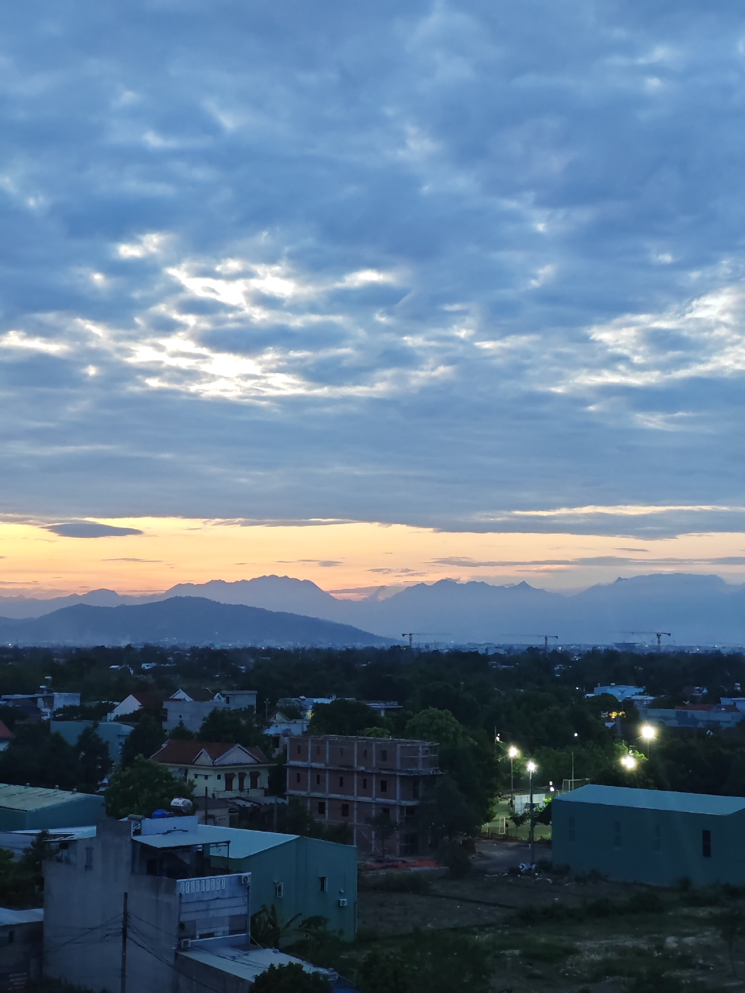 The image captures a twilight scene over a residential area in Đà Nẵng, Vietnam, likely taken from an elevated viewpoint. The sky is dominated by a gradient of blues and grays, with scattered clouds tinged with the soft orange and yellow hues of a setting or rising sun. In the distance, a range of mountains forms a hazy silhouette against the horizon. Below the sky, the cityscape unfolds, characterized by a dense collection of buildings, ranging from single-family homes with tiled roofs to multi-story structures under construction. Lush green trees are interspersed throughout the urban landscape, softening the edges of the built environment. Several streetlights illuminate parts of the scene, suggesting it is either dawn or dusk. Construction cranes are visible in the far distance, indicating ongoing development. The overall impression is one of a developing urban area at the edge of natural landscape, under a vast, cloudy sky.