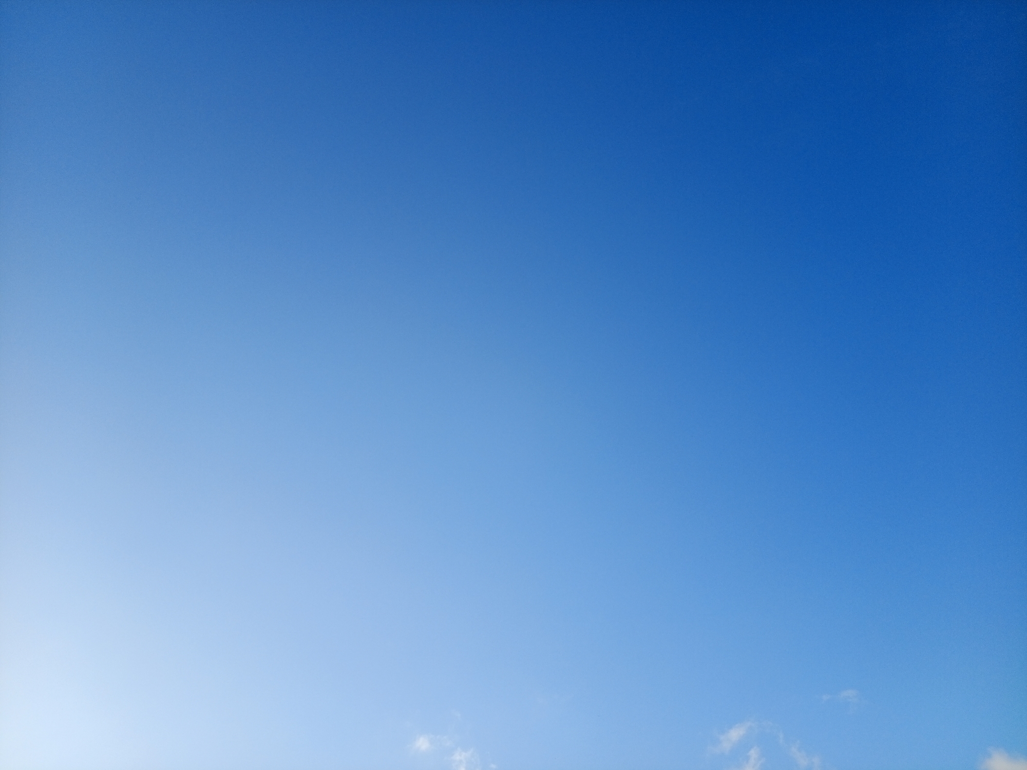 The image depicts a clear blue sky. The color gradient transitions from a lighter, almost white hue in the bottom left corner to a deeper blue towards the top right. A few small, wispy clouds are visible in the bottom right corner. The location and time are unknown.