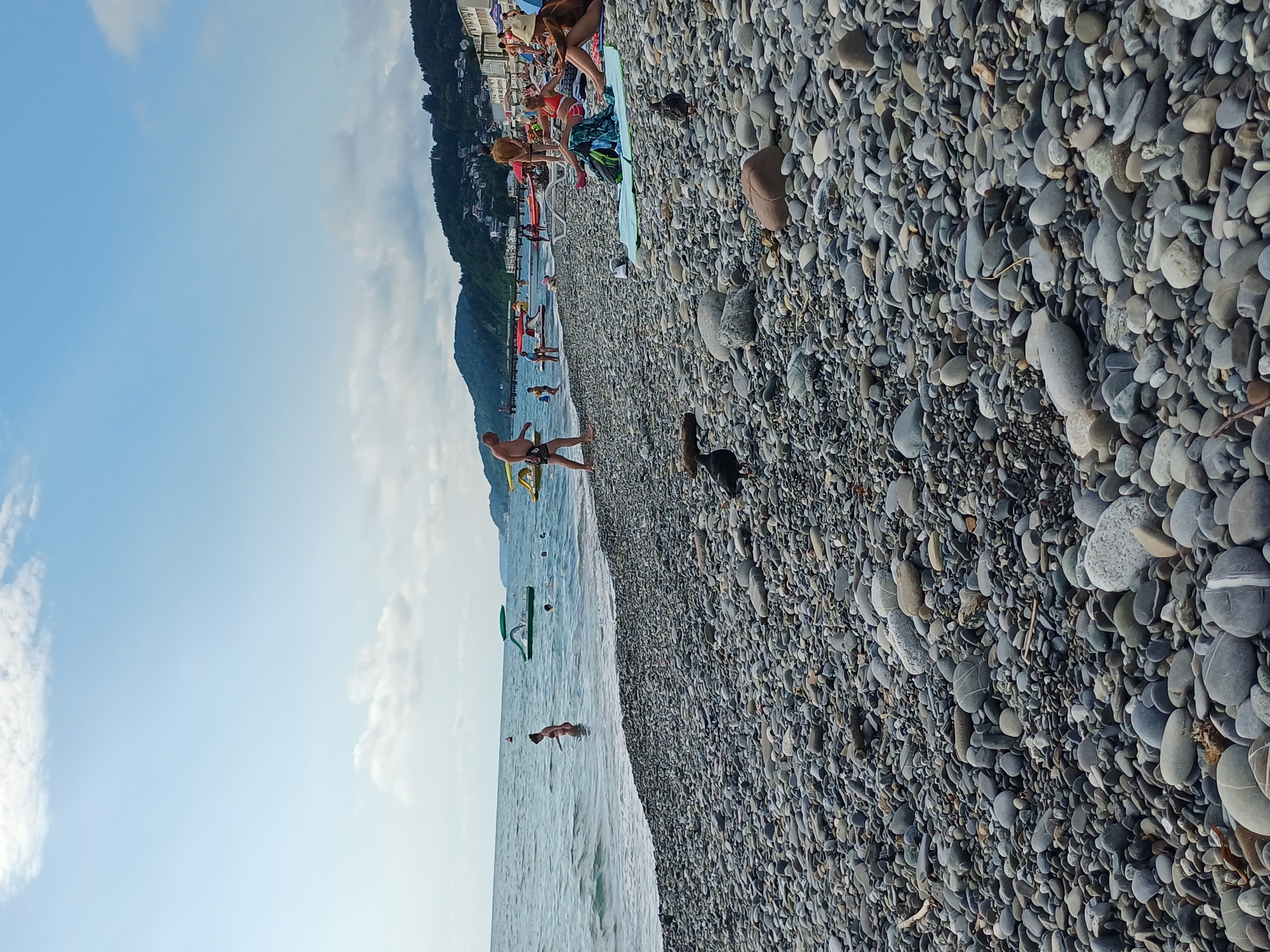 The image depicts a lively scene on a pebble beach in Sochi, Russia. People are scattered across the beach and in the shallow water, enjoying a sunny day. On the beach, some individuals are sunbathing or sitting, while others are walking or playing. In the water, several people are swimming, wading, or engaging in water activities. A few colorful beach umbrellas and inflatable toys add to the vibrant atmosphere.
The background shows a lush green hillside with buildings dotting its slopes, characteristic of the coastal landscape of Sochi. The sea is calm with gentle waves lapping against the shore. The sky is a clear blue with scattered white clouds, suggesting fair weather. The time of day appears to be midday or early afternoon, given the bright sunlight casting shadows.
A man in black shorts is standing near the water's edge, looking towards the sea. Further back, a person in a red swimsuit is visible. There is also a dark-colored dog on the beach, adding a touch of domesticity to the scene. The abundance of smooth, gray pebbles covering the beach is a prominent feature of the landscape. The overall mood of the image is relaxed and leisurely, capturing the essence of a summer day at the beach. No visible text is present in the image.