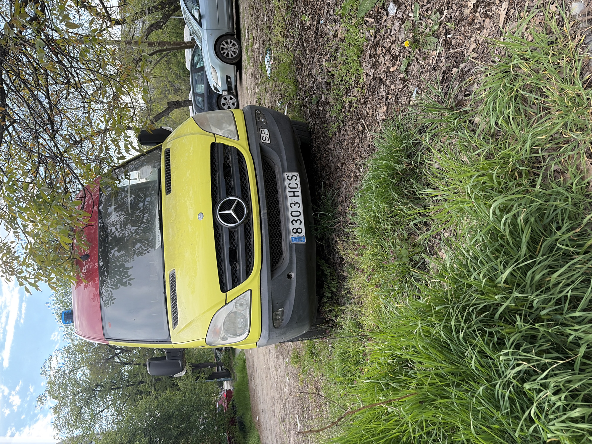 The image captures a yellow and red Mercedes-Benz Sprinter van, likely an emergency vehicle such as an ambulance, parked outdoors in what appears to be an unpaved area. The van's front is primarily yellow, featuring the distinctive Mercedes-Benz emblem on its black grille, flanked by clear headlights. A black bumper runs across the lower front. Above the windshield, the roof is red, and a blue emergency light is visible on the left side of the roof.
A white license plate is prominently displayed on the front bumper, bearing the alphanumeric sequence "8303 HCS". The "BG" country code is visible on the left side of the plate, confirming the location as Bulgaria. A small square sticker with "SP" is also affixed to the black bumper.
To the right of the van, the foreground is dominated by lush green grass. The ground immediately around the vehicle and extending into the middle distance is a mix of dirt, dry leaves, and sparse vegetation. In the background, beyond the yellow van, other vehicles are parked, including a light blue or silver sedan and a darker car, suggesting a parking area.
The scene is set outdoors during the daytime under a partly cloudy blue sky, visible through the branches of trees with green leaves that frame the upper left portion of the image. The overall environment appears somewhat rustic or undeveloped, characteristic of peripheral areas in locations like Plovdiv, Bulgaria. No people or specific activities are visible, focusing solely on the parked vehicles and their natural surroundings.