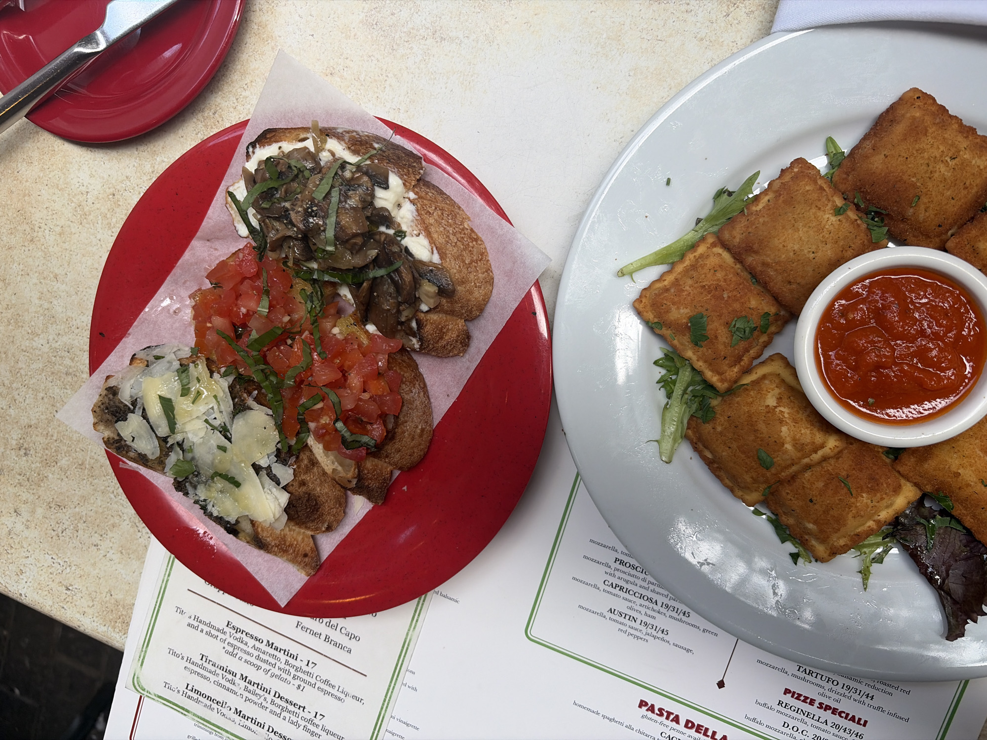 The image shows a table setting at an Italian restaurant, likely in Austin, Texas, based on the "AUSTIN 19/31/45" menu item visible in the lower right. Two plates are prominently displayed. One is a red plate with three pieces of bruschetta topped with various ingredients, including tomatoes, mushrooms, and cheese. The other is a white plate with fried ravioli served with a dipping sauce in a small bowl. The restaurant menu is partially visible in the foreground, with items like "Espresso Martini," "Tiramisu Martini Dessert," and "Limoncello Martini Dessert" listed. The menu also includes pizza and pasta dishes. A silver fork is on a smaller red plate in the upper left corner.