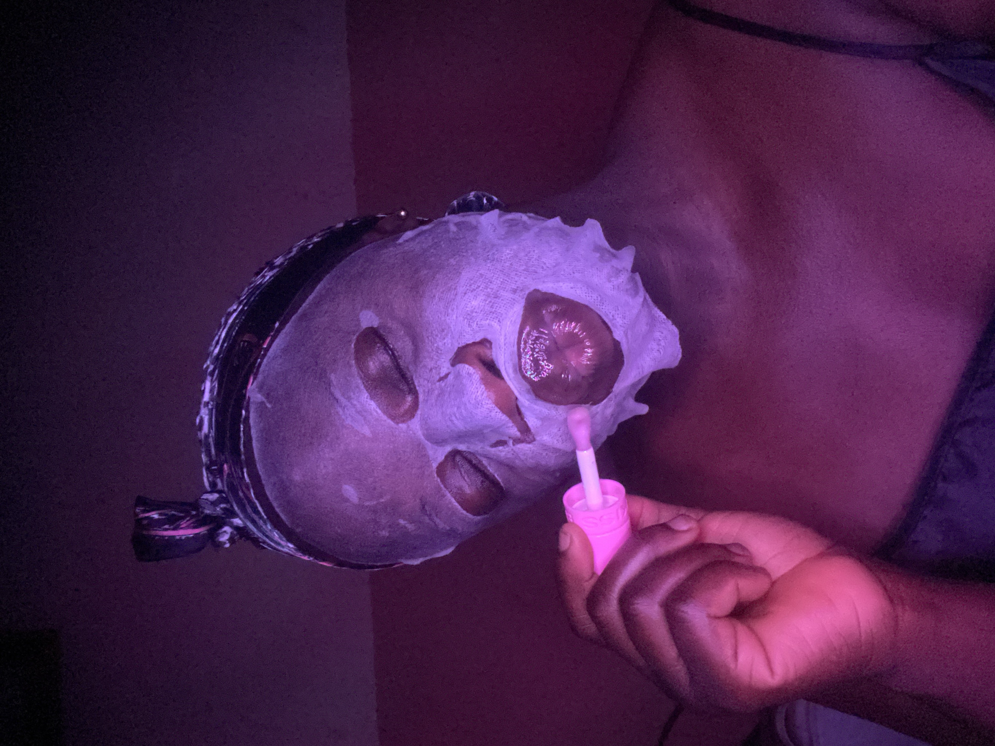 The image depicts a close-up of a person, likely in Maigizo, Nigeria, engaged in a self-care routine within an indoor setting. The scene is illuminated by strong, artificial purple and pink lighting, creating a distinct ambiance. The subject, who has dark skin, is wearing a white or light-colored sheet mask on their face, which covers their forehead, cheeks, and chin, with cut-outs for the eyes, nose, and mouth.
In the lower right of the frame, the person's right hand is visible, holding a bright pink lip gloss tube with its applicator extended. The applicator is positioned near the opening for the mouth in the sheet mask. Part of the person's braided hair and a dark head covering are visible on the left side of their head. A dark strap, possibly from clothing, can be seen over the person's shoulder and upper chest. The background is dark and indistinct, suggesting a wall or a shadowed area, with no specific details discernible. The absence of natural light indicates that the activity is taking place in the evening or night, or in a specially lit room. There is no visible text within the image.