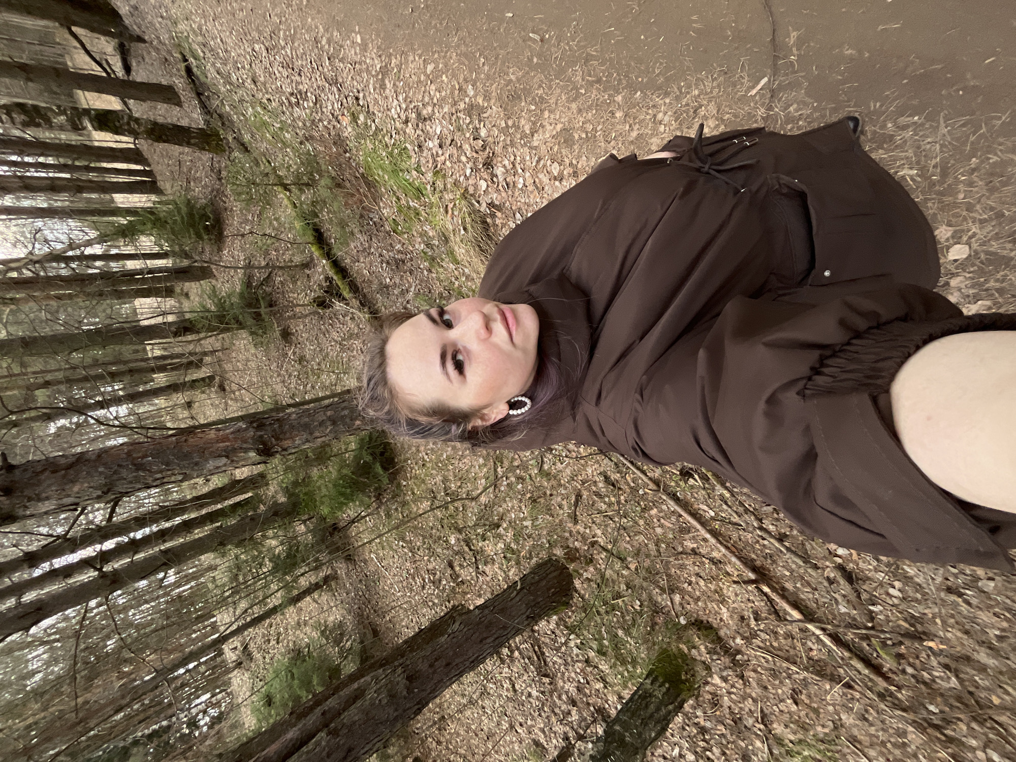 A young woman is featured in a slightly low-angle, upright portrait taken in a wooded area in Chaikovsky, Russia. She looks directly at the camera with a neutral to slightly pleasant expression. Her light-colored hair, possibly with a subtle purple tint, frames her face. She is wearing a dark brown, padded jacket or coat, suitable for cooler weather. A distinctive earring, appearing to be a white, possibly pearl-studded, hoop, is visible on her left ear.
The setting is a forest or wooded environment. The ground beneath her is covered with a thick layer of dry, fallen leaves and some sparse green foliage, suggesting late autumn, winter, or early spring. Tall trees, including both coniferous and leafless deciduous varieties, rise vertically in the background, creating a dense backdrop. The lighting appears soft and diffused, indicating an overcast day or deep shade within the forest. No visible text is present in the image.