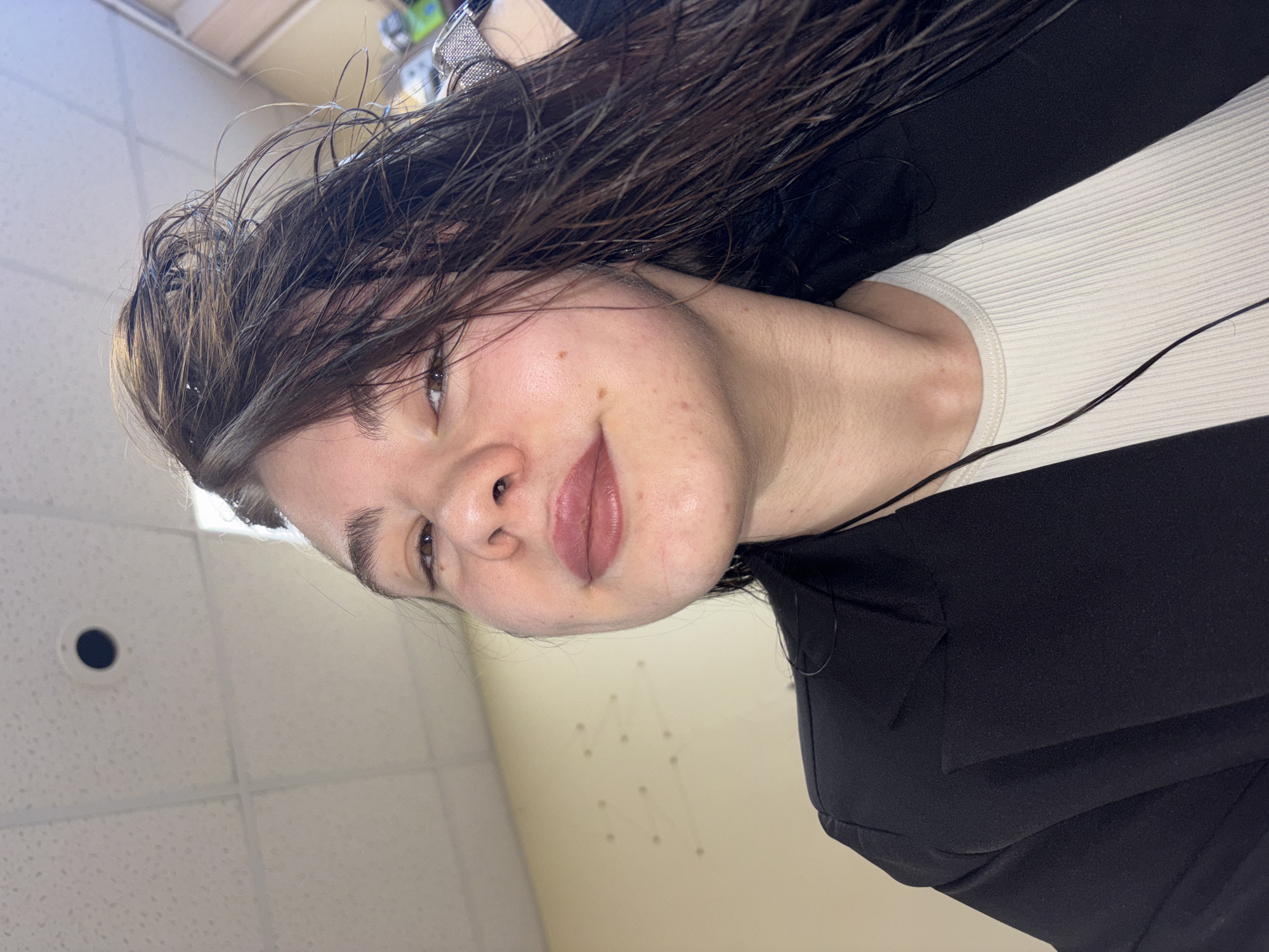 A woman with wet, dark brown hair and fair skin poses for a selfie. She is wearing a white ribbed top under a black blazer. The background is a drop ceiling with square panels, a wall with a pattern of small dots, and what appears to be part of a shelf. The location is not discernible to be Perm, Russia.